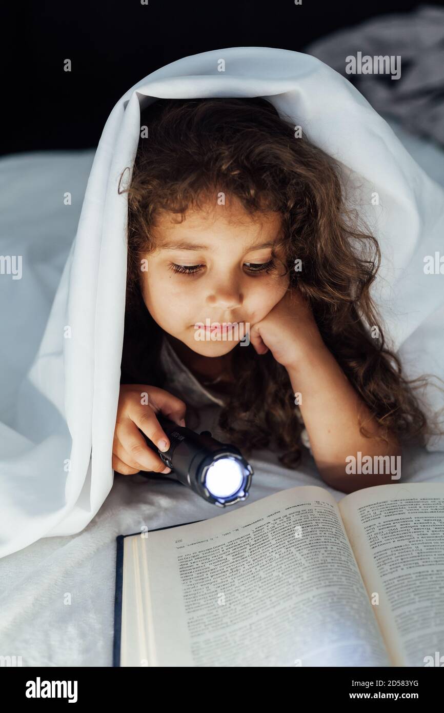Schönes kleines Mädchen liest ein Buch mit einer Laterne an Nacht unter einer Decke Stockfoto