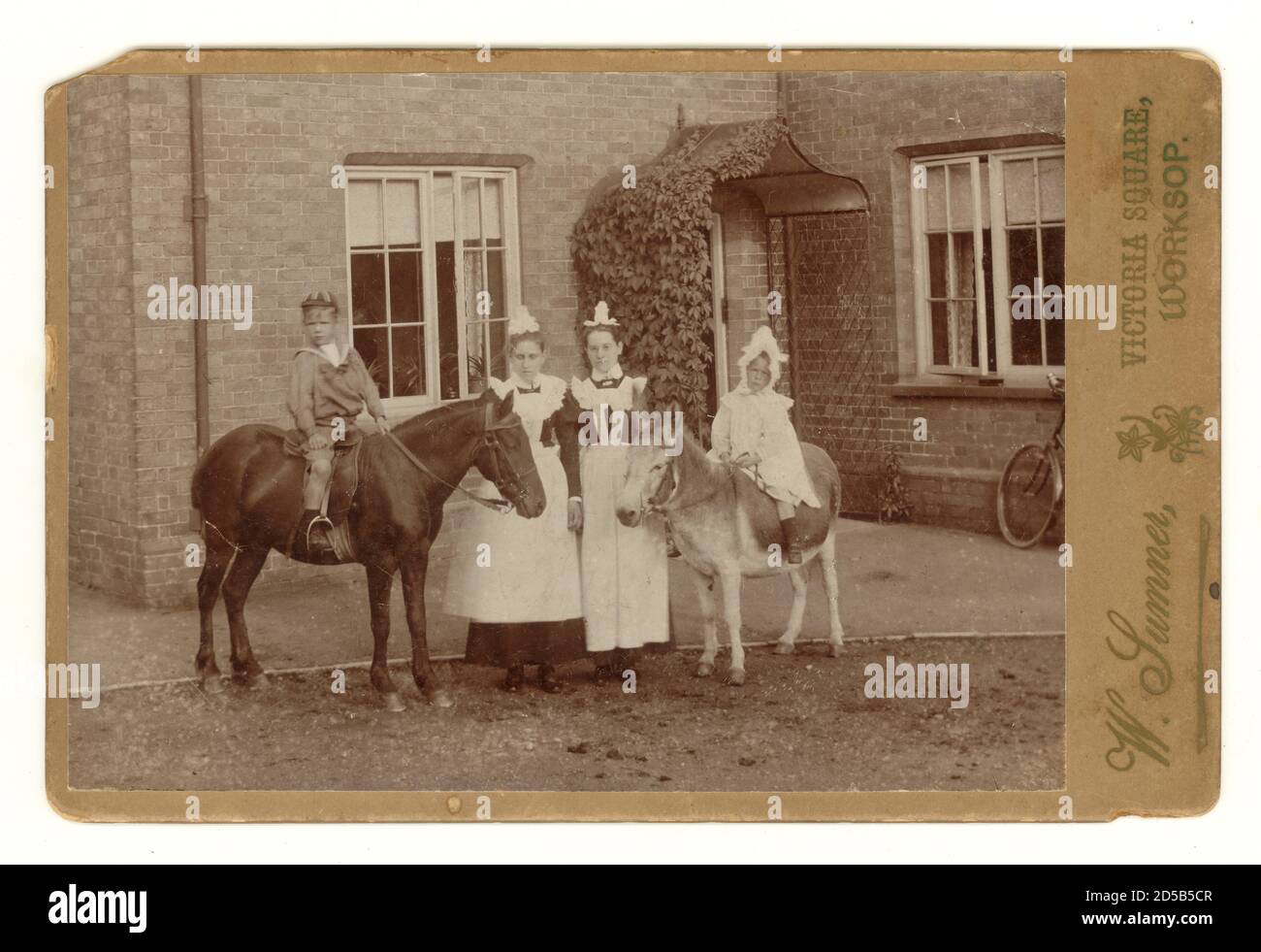 Originale typisch charmante viktorianische Kabinettkarte aus den 1890er Jahren mit kleinen Kindern auf einem Esel und einem Pony vor einem Landhaus mit den Stubenmädchen, viktorianischen Dienstmädchen, viktorianischen Dienstmädchen, Worksop, Nottinghamshire, England, Großbritannien um 1894 Stockfoto