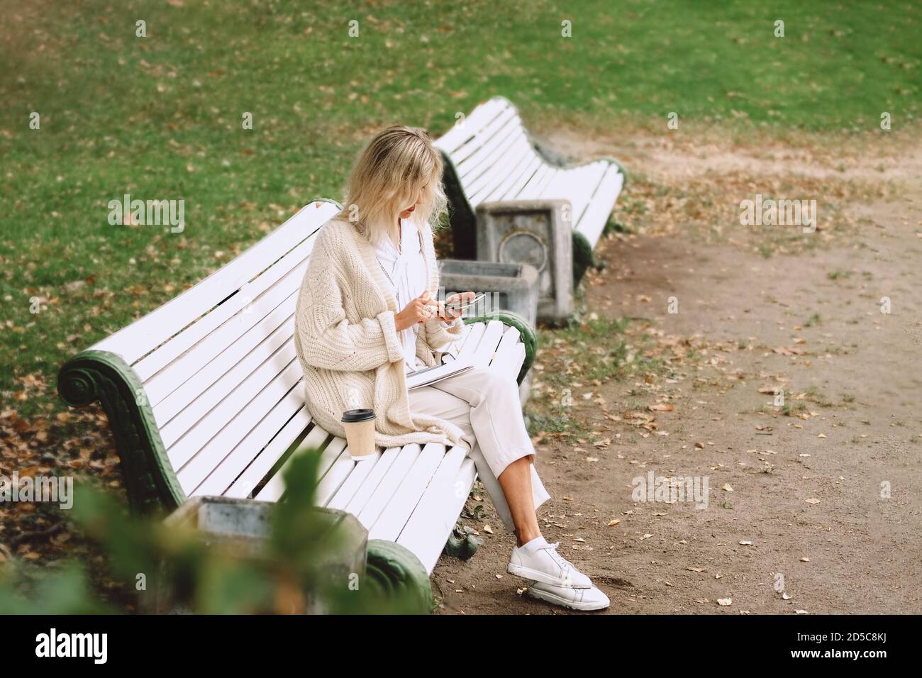 Junge Geschäftsfrau sitzt auf der Bank im Herbstpark und spricht über das Telefon Notizen machen. Kaffeepause im Freien. Stockfoto