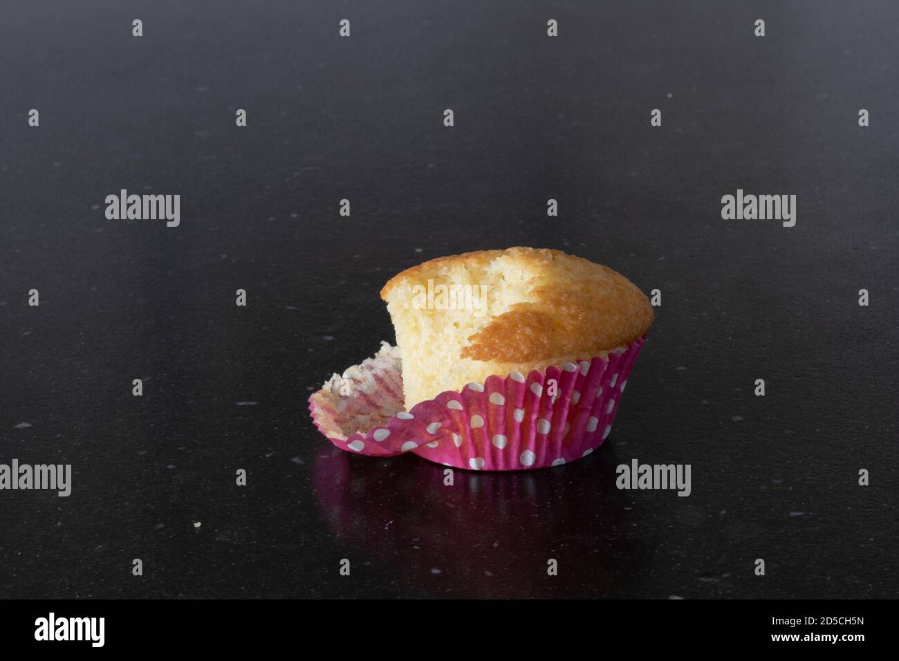 Nehmen einen Biss von einem Plein Cupcake, brechen eine Diät. Rosa Tasse mit weißen Punkten. Dunkelgrauer Hintergrund, Küchentisch Stockfoto