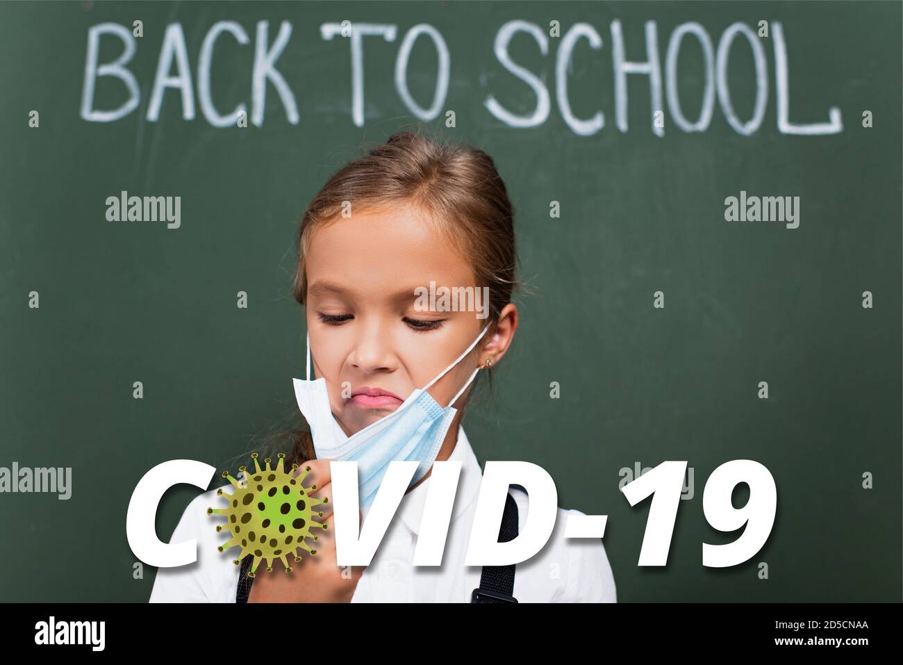 Selektiver Fokus der unzufrieden Schulmädchen berühren Schutzmaske in der Nähe Kreidetafel Und covid-19 Schriftzug im Klassenzimmer Stockfoto