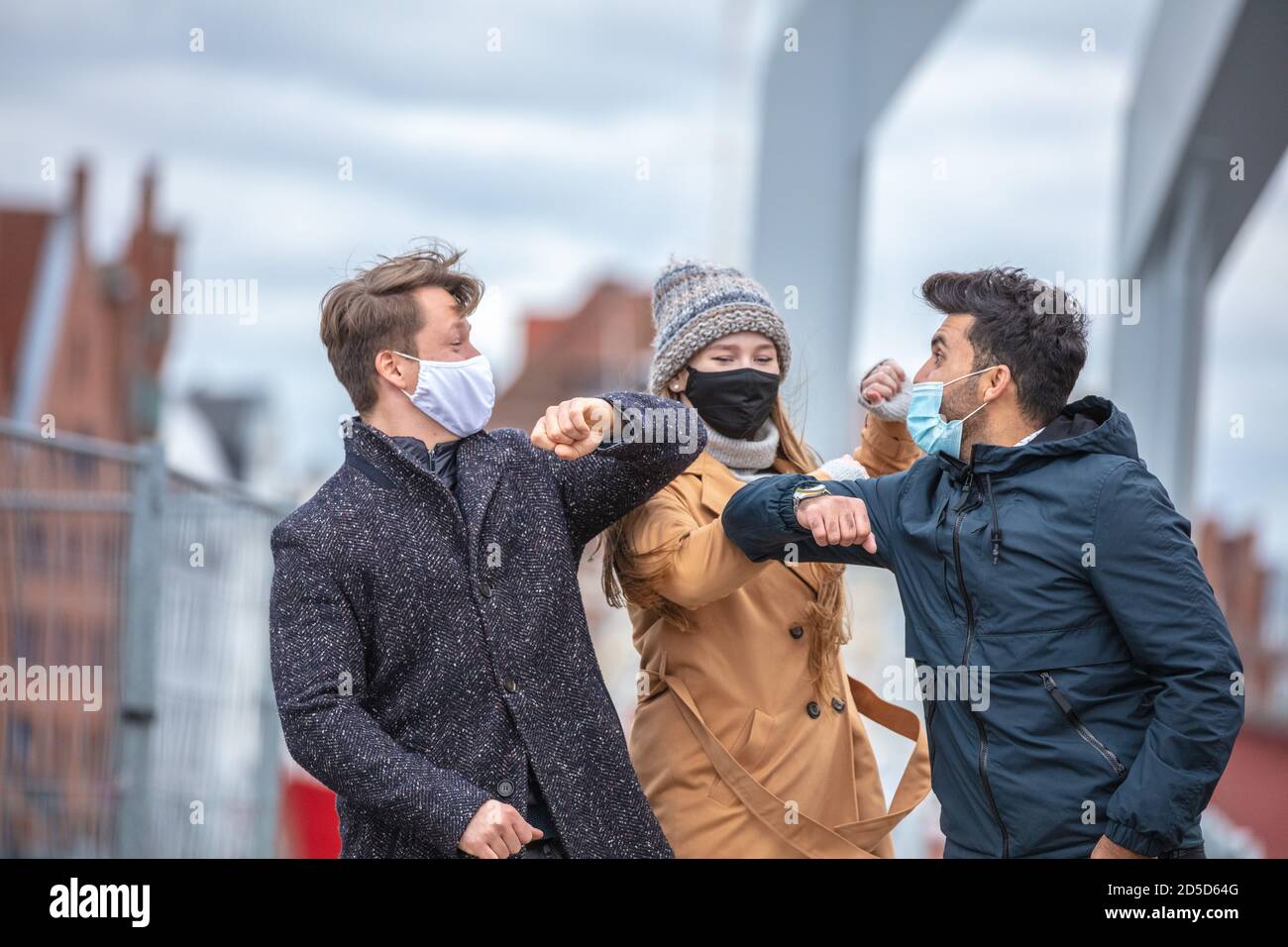 Zwei Männer und eine Frau grüßen sich mit dem Ellenbogen. Corona-Zeit mit alltäglichen Masken in der kalten Jahreszeit in der Stadt. Stockfoto