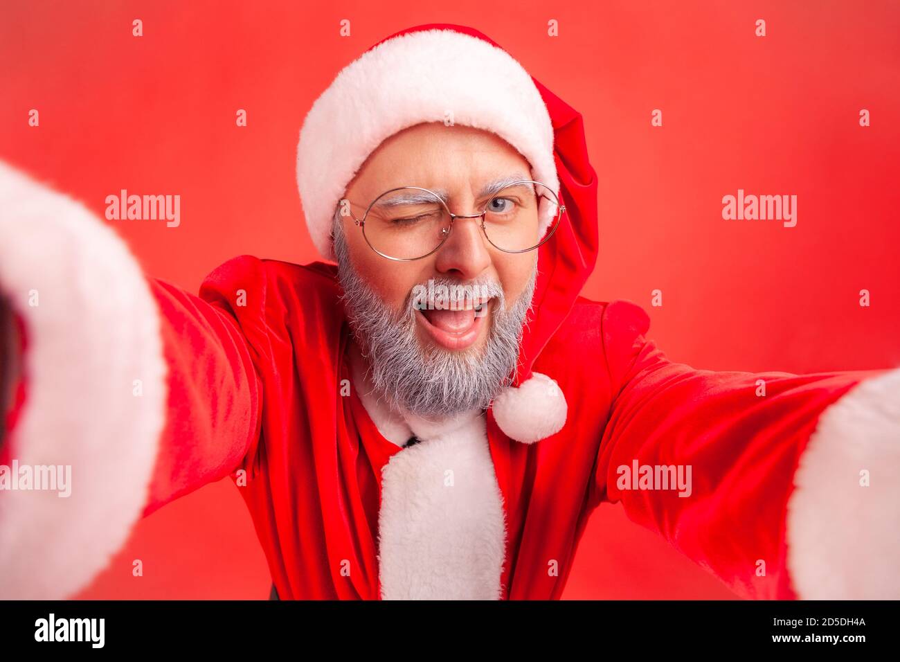 Nahaufnahme lächelnd positiver graubärtiger Mann in Brillen und weihnachtsmann Kostüm, der Kamera anschaut und zwinkert, Selfie macht. Innenaufnahme des Studios isola Stockfoto