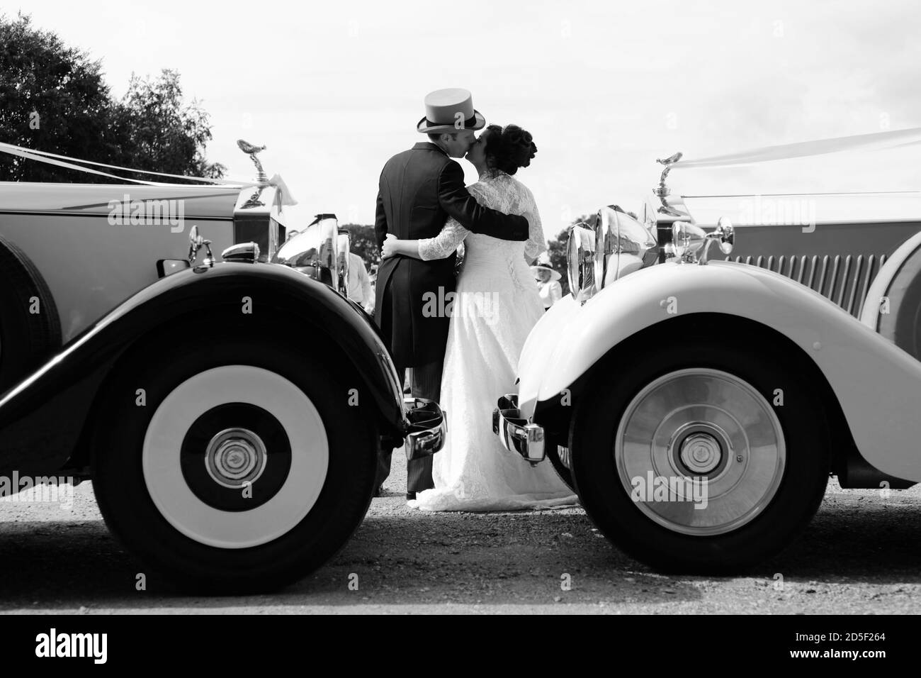 Braut in weißem Hochzeitskleid und Bräutigam in grauem Hut und Schwänze küssen zwischen Rolls Royce Hochzeitsautos. Braut & Bräutigam küssen am Hochzeitstag. Stockfoto