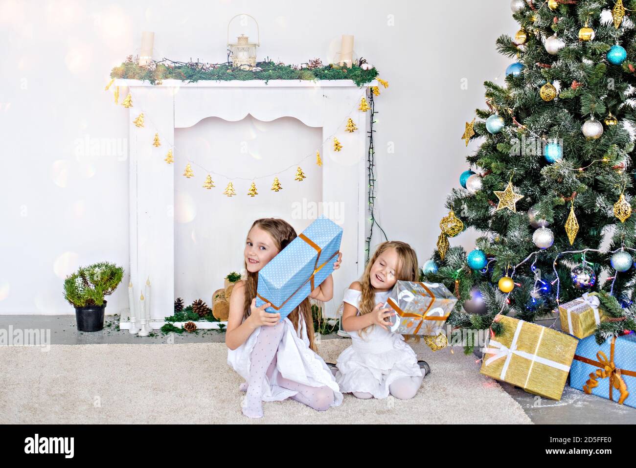 Zwei kleine blonde Mädchen in weißen Kleidern spielen am Kamin und am Weihnachtsbaum mit Kisten. Schwestern haben Spaß, umarmen, lachen, entfalten Geschenke Stockfoto