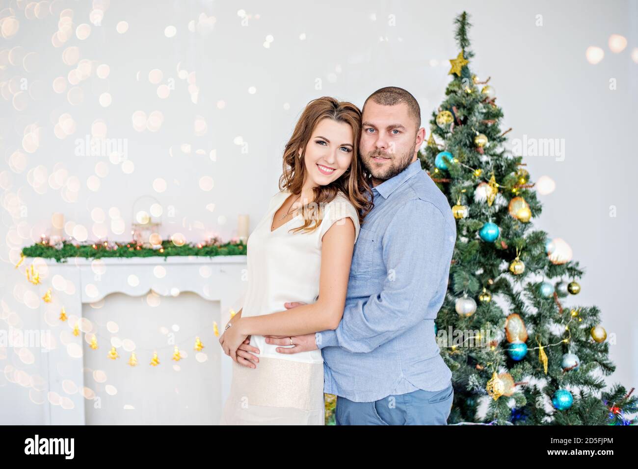 Nahaufnahme eines verliebten Paares vor dem Hintergrund eines Bokeh eines Weihnachtsbaums. Ein junger Mann mit einem Bart umarmt eine schöne Frau in einem Stockfoto