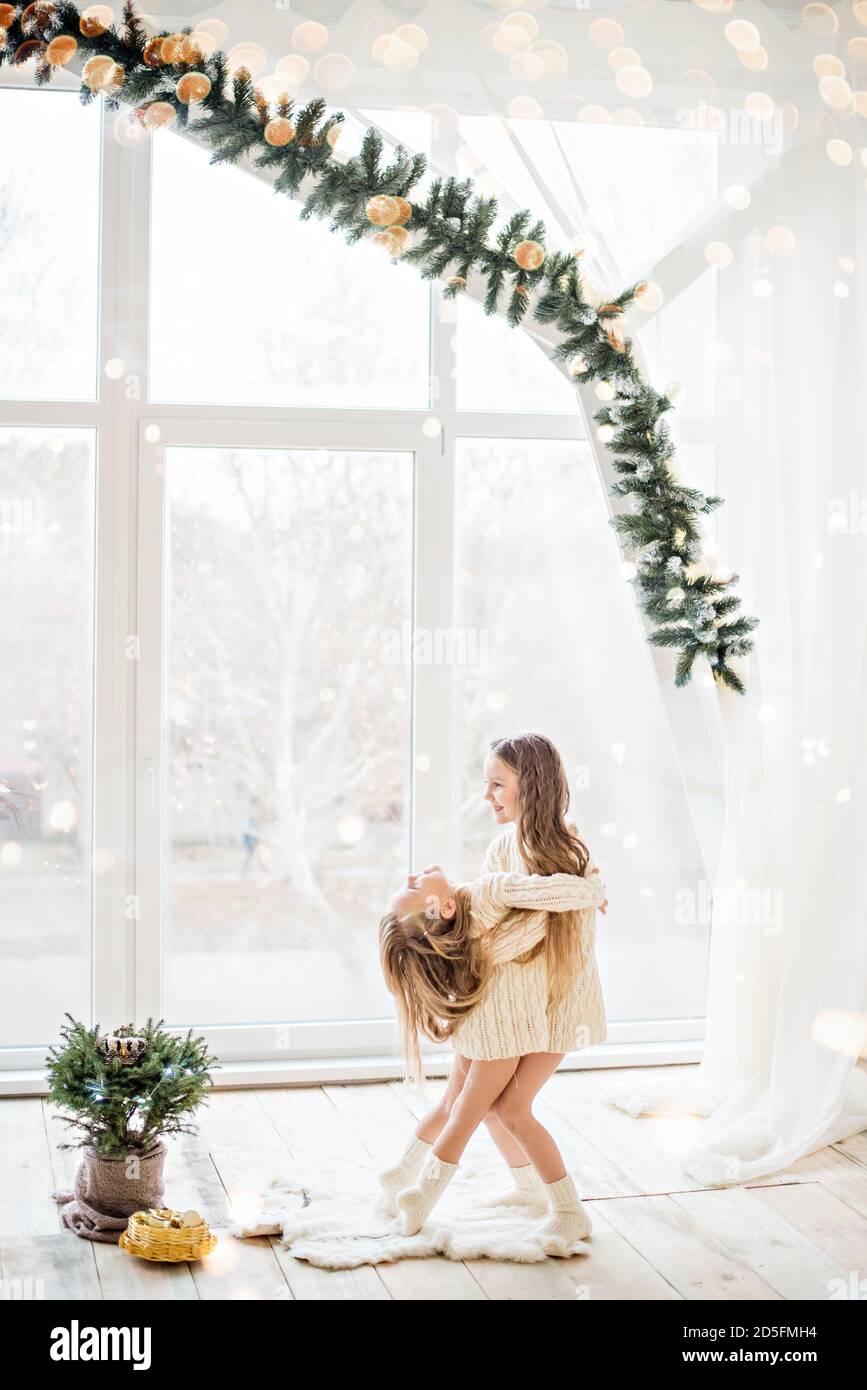 Zwei Freundinnen der blonden Schwester in gestrickten weißen Pullovern Und die Socken spielen am Panoramafenster mit Weihnachten Bäume in Erwartung Stockfoto