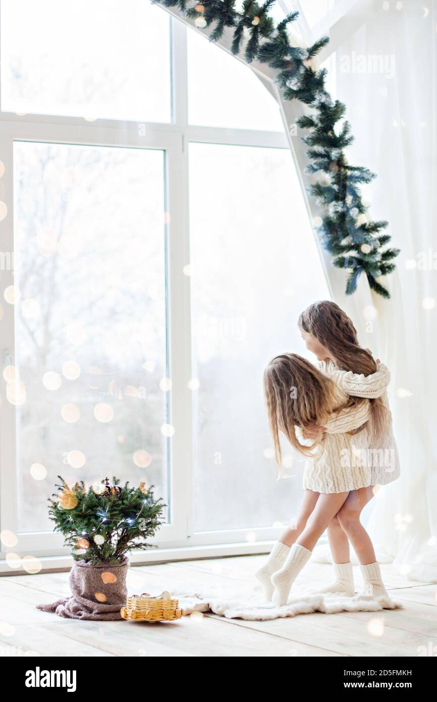 Zwei Freundinnen der blonden Schwester in gestrickten weißen Pullovern Und die Socken spielen am Panoramafenster mit Weihnachten Bäume in Erwartung Stockfoto