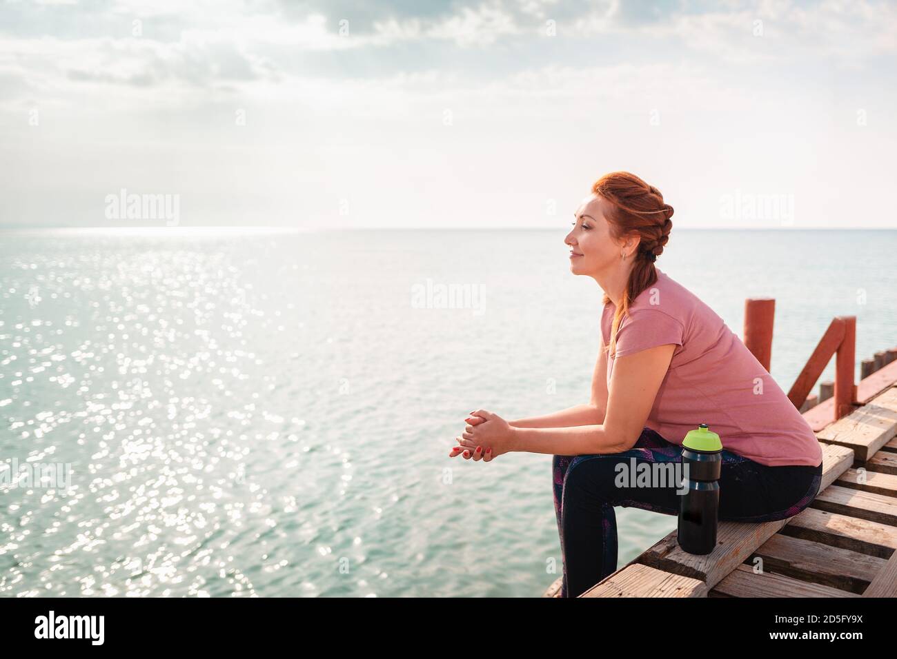 Meer und Freiheit. Erwachsene Frau in Sportkleidung sitzt auf dem Pier. Seitenansicht. Speicherplatz kopieren. Stockfoto