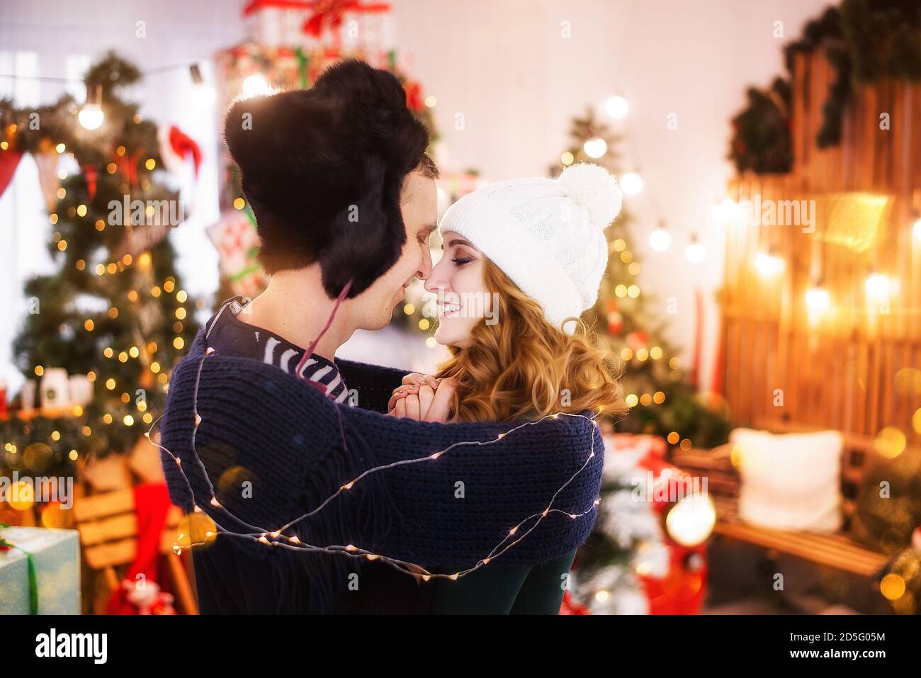Ein Nahaufnahme-Porträt eines verliebten jungen Paares vor dem Hintergrund von Weihnachtsbäumen mit Girlanden. Ein junger Mann in einem Hut mit Ohrenklappen wärmt Stockfoto