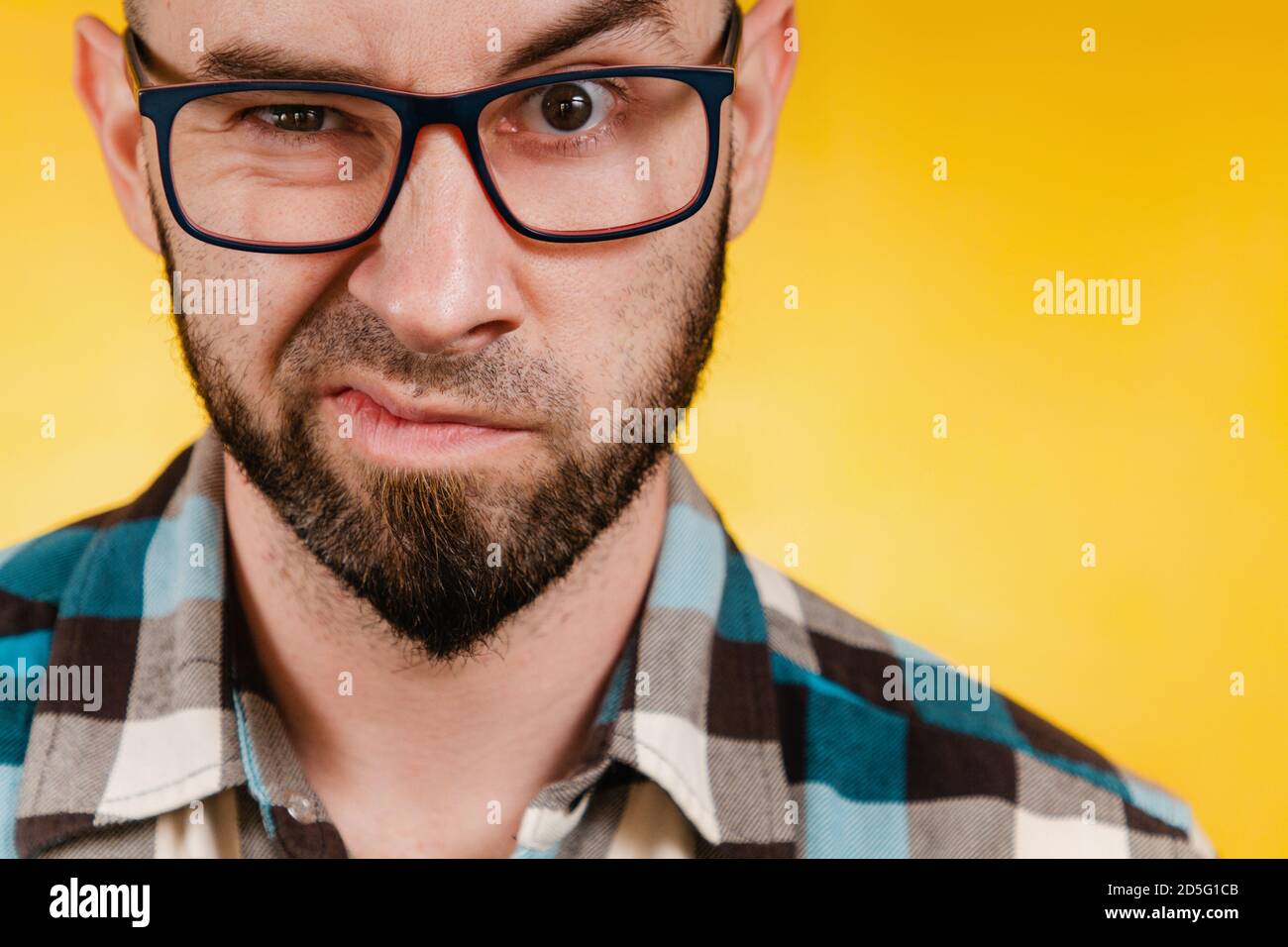 Menschen und Emotionen. Ein bärtiger Mann mit Brille und einem blau karierten Hemd drückt seine Abneigung aus. Gelber Hintergrund. Nahaufnahme. Stockfoto