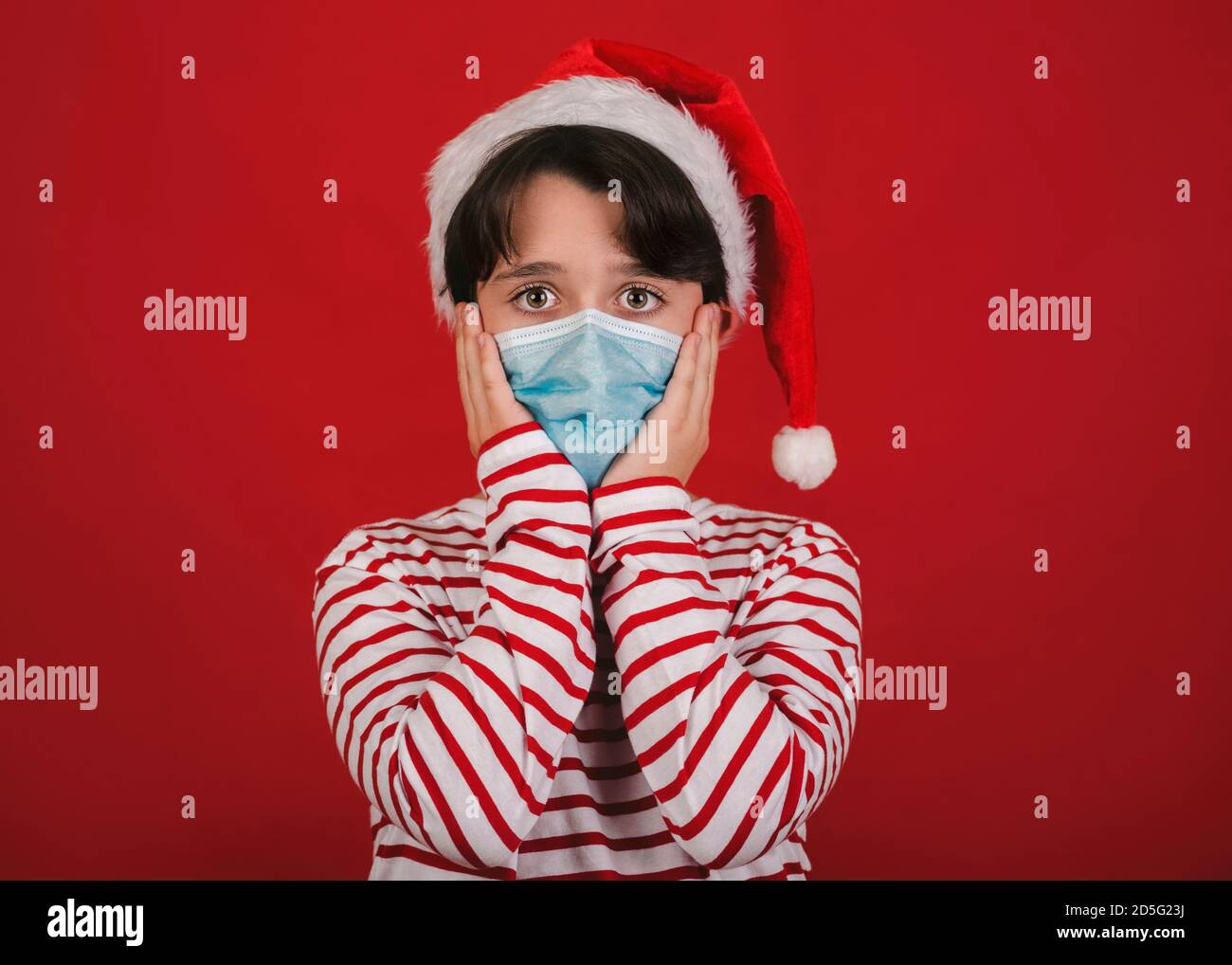 Frohe Weihnachten, lustiges Kind mit medizinischer Maske trägt Weihnachtsmann Hut Auf rotem Hintergrund Stockfoto