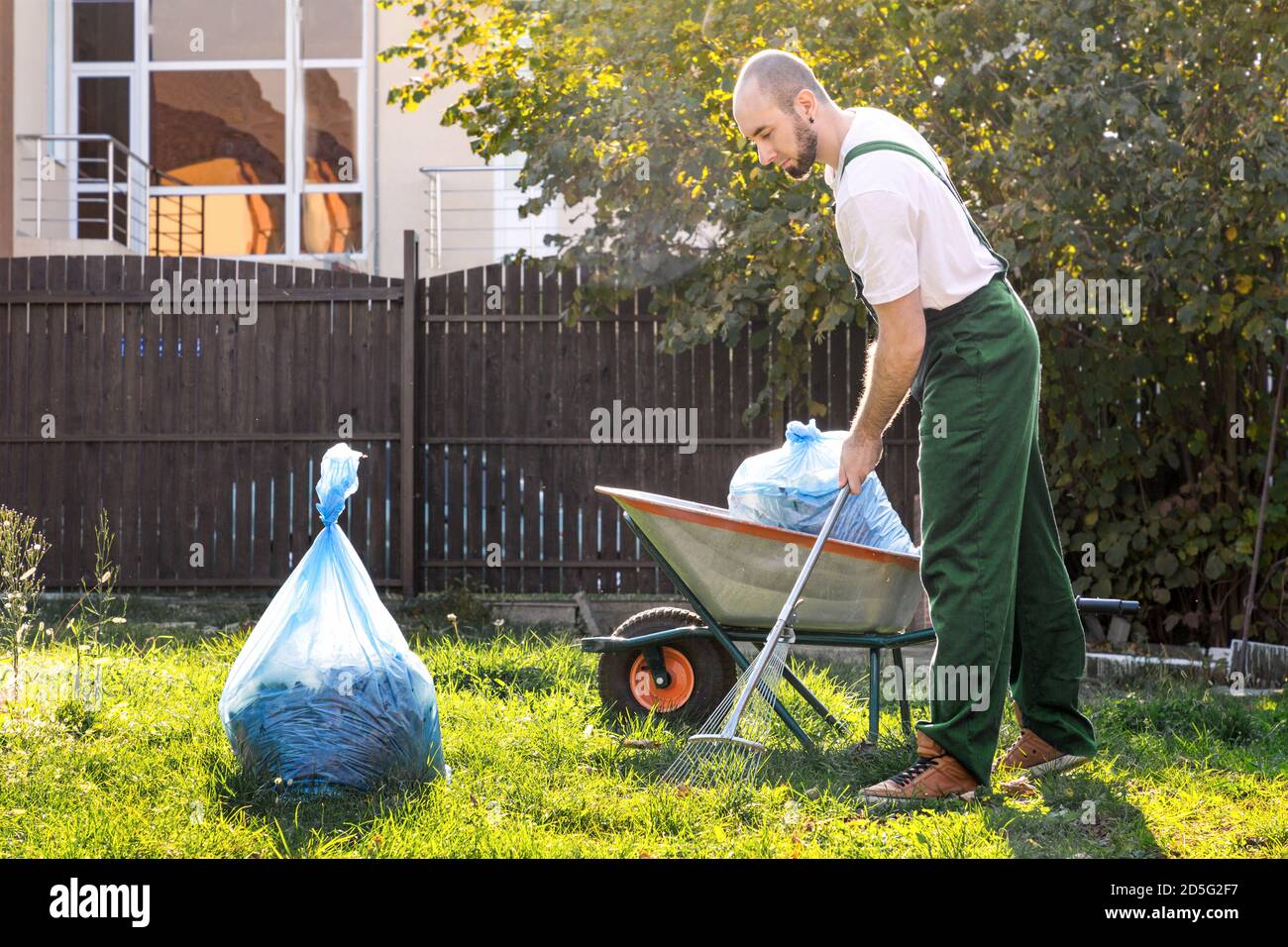 Der Gärtner in der grünen Uniform reinigt den Hof.auf dem Gras ist ein Wagen mit Kompost und einer Müllpackung. Stockfoto