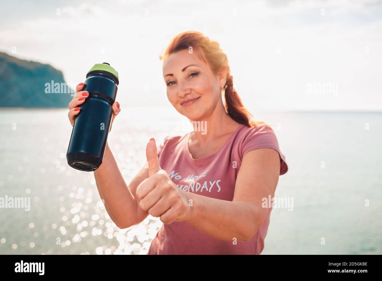 Portrait einer Frau in Sportkleidung, die einen Shaker hält und einen Daumen nach oben gibt. Meer im Hintergrund. Das Konzept der Sporternährung. Stockfoto