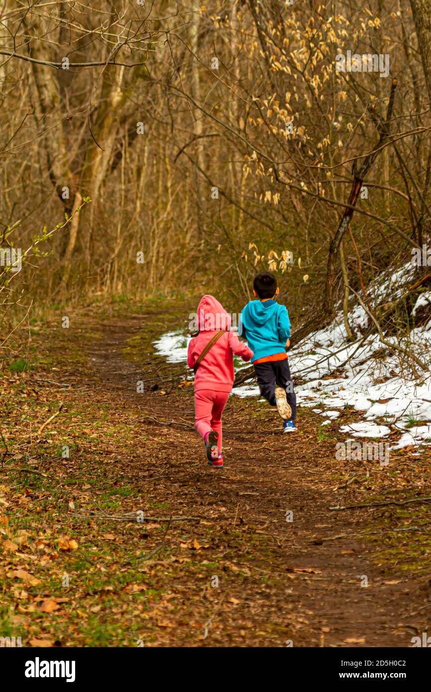 Ein Junge und ein Mädchen laufen auf einem Wanderweg im Wald. Die Blätter sind gefallen und der Weg ist mit Blättern bedeckt. Diese energischen Kinder tragen wint Stockfoto