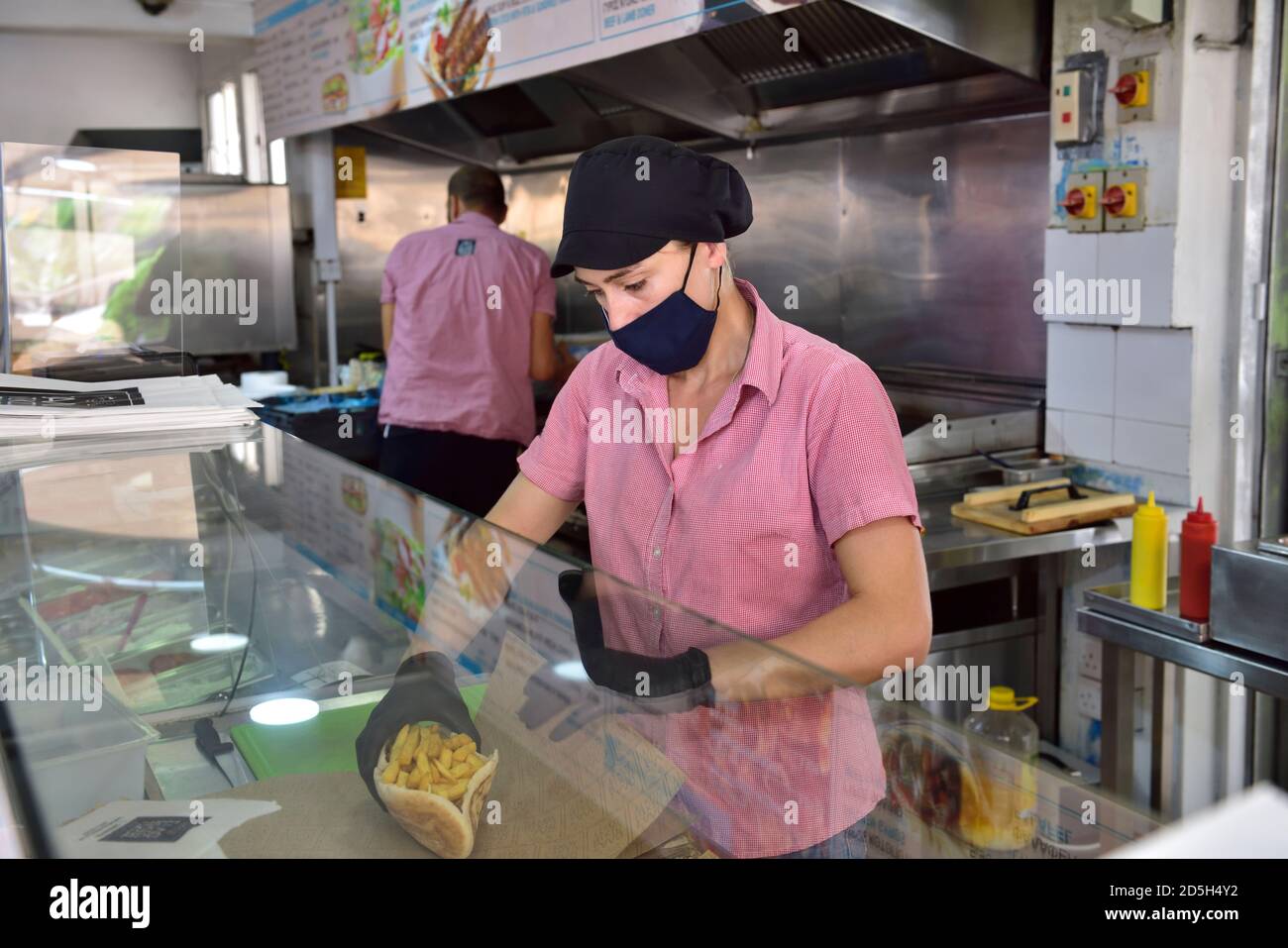 Junge Frau, die Essen (Gyros, Kebab) für einen Kunden vorbereitet, während sie eine Gesichtsmaske und Handschuhe trägt, aufgrund von Covid-19 Vorsichtsmaßnahmen Stockfoto