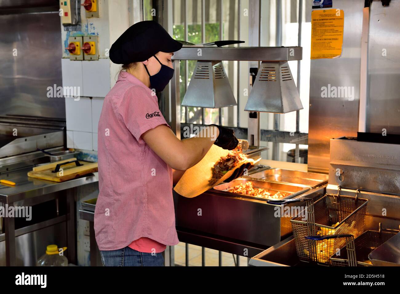 Junge Frau, die Essen (Gyros, Kebab) für einen Kunden vorbereitet, während sie eine Gesichtsmaske und Handschuhe trägt, aufgrund von Covid-19 Vorsichtsmaßnahmen Stockfoto