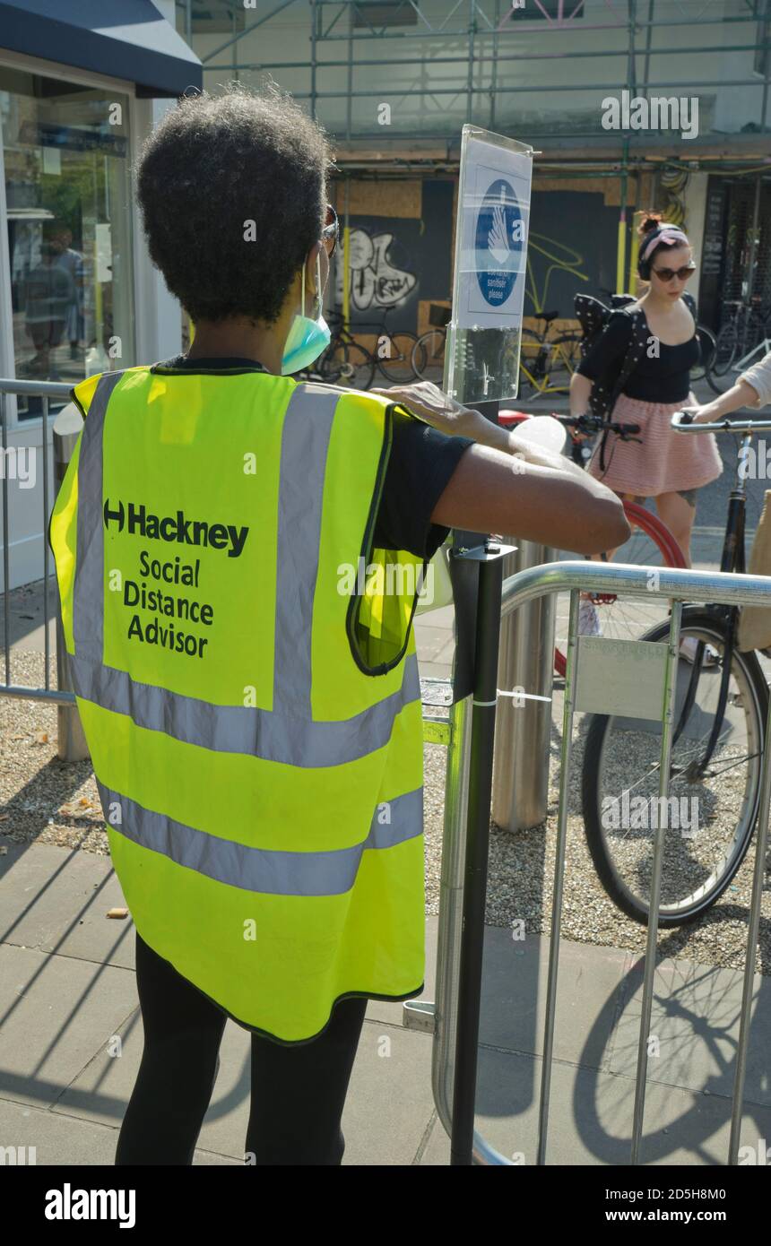 Soziale Distanzberater helfen Besuchern des Broadway Market, mit Straßenschildern, die darauf hinweisen, dass die soziale Distanz aufgrund der Coronavirus / Covid-19 Pandemie in Hackney, London, England, Großbritannien, geschützt ist Stockfoto