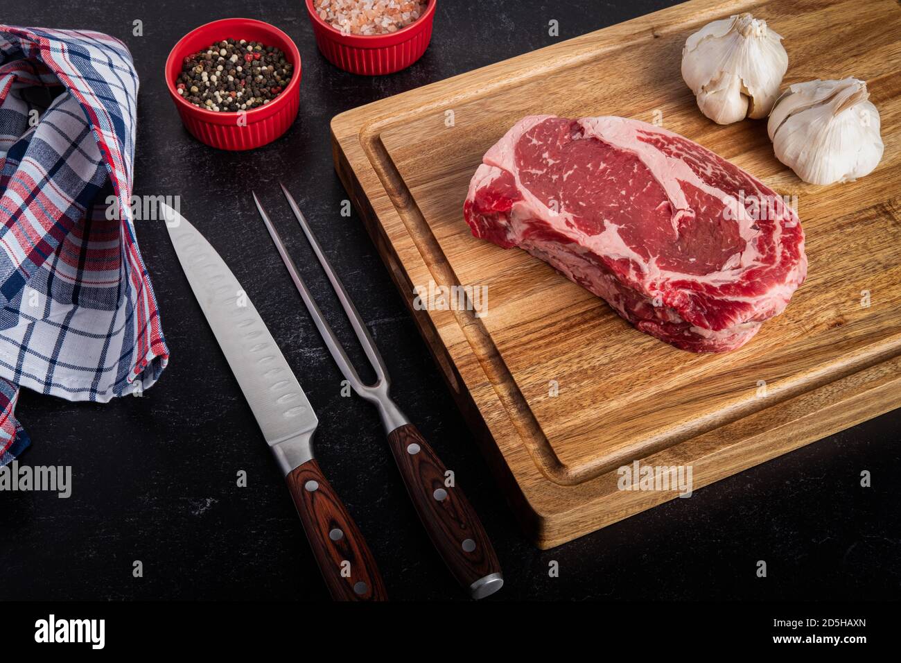Foto von rohem Ribeye Steak ohne Knochen mit Meersalz, Pfeffer, Knoblauch auf einem Schneidebrett mit einem Schnitzmesser und Gabel-Set Stockfoto