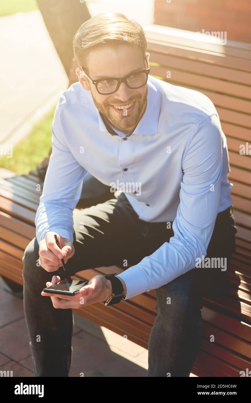 Vertikale Aufnahme eines jungen fröhlichen Geschäftsmann in blauem Hemd Und Brillen mit Smartphone und lächeln, während Sie auf dem sitzen Bank im Freien auf einem Stockfoto