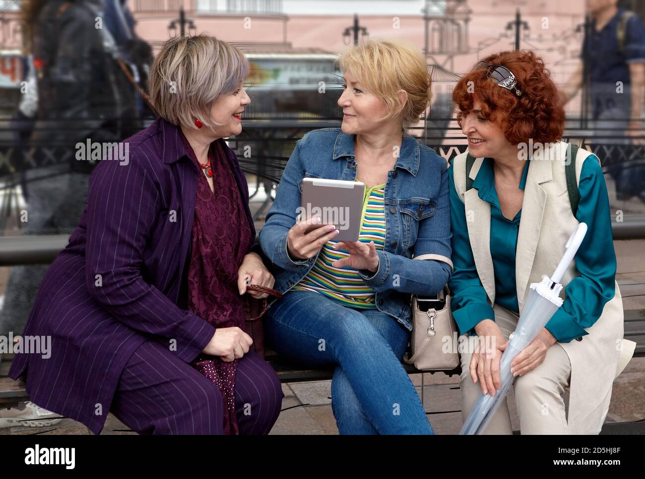 Drei lächelnde europäische Frauen mittleren Alters kommunizieren an einer Bushaltestelle auf einer Bank sitzend. Stockfoto