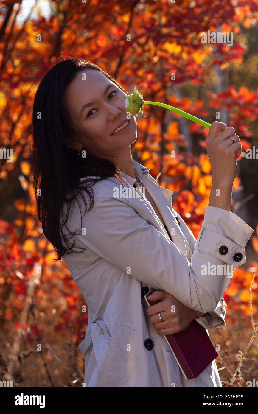 Porträt von lächelnden asiatischen jungen Frau mit Buch in ihr Hand und Blume an ihrem Gesicht im Herbstwald Stockfoto