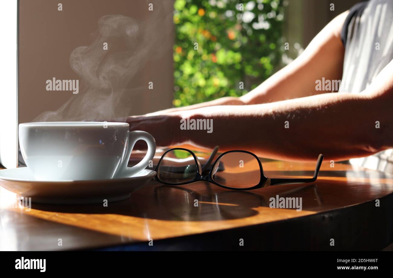 Frau tippt auf dem Laptop mit einer Brille und einem dampfenden heißen Getränk auf dem Schreibtisch. Arbeiten im Heimbüro. Stockfoto
