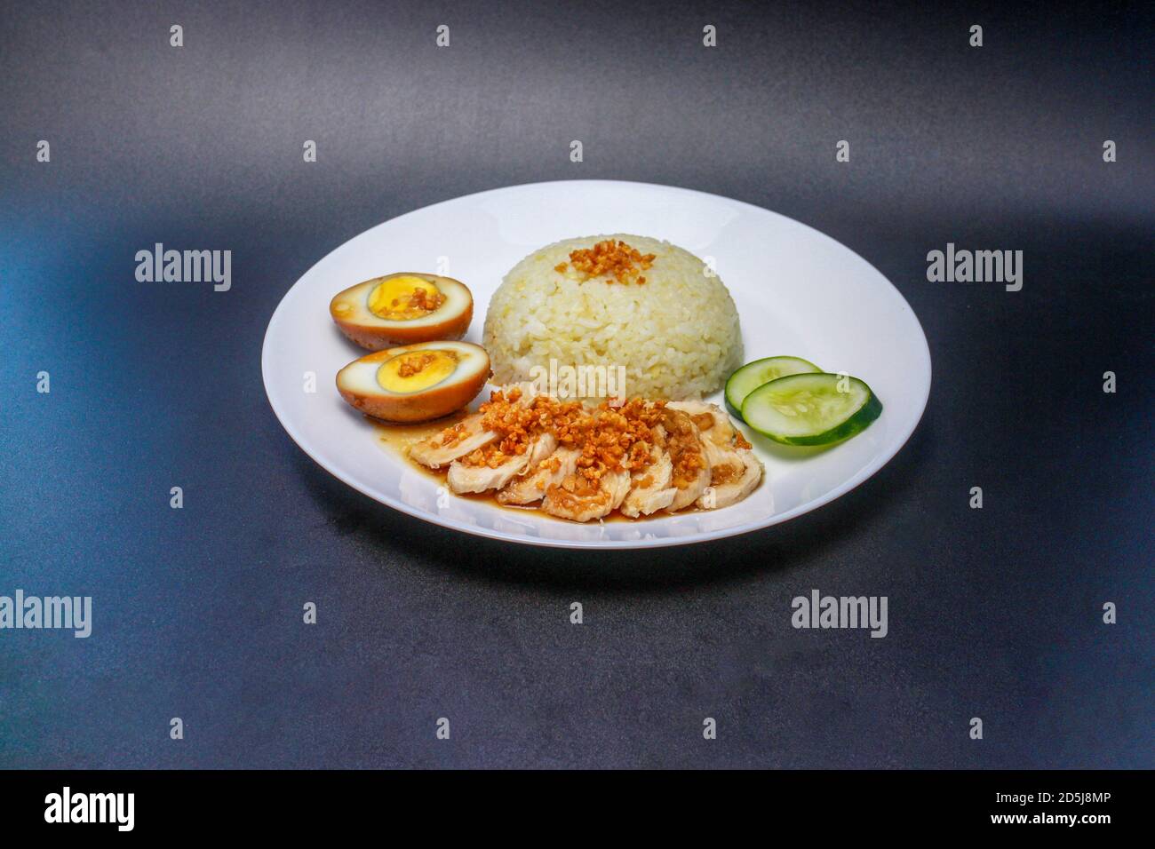 Hainanese Hühnerreis oder gedämpfter Hühnerreis mit süßem Sojasauce-Ei und Gurkenscheiben. Serviert auf einem weißen Teller. Stockfoto