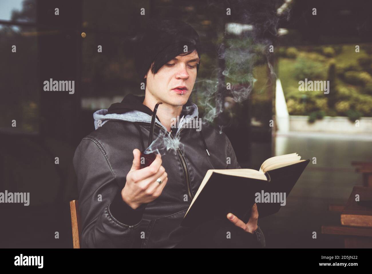 Junger schwarzhaariger Mann, der im Café im Freien ein Buch durchbohrt und Tabakpfeife raucht. Stockfoto