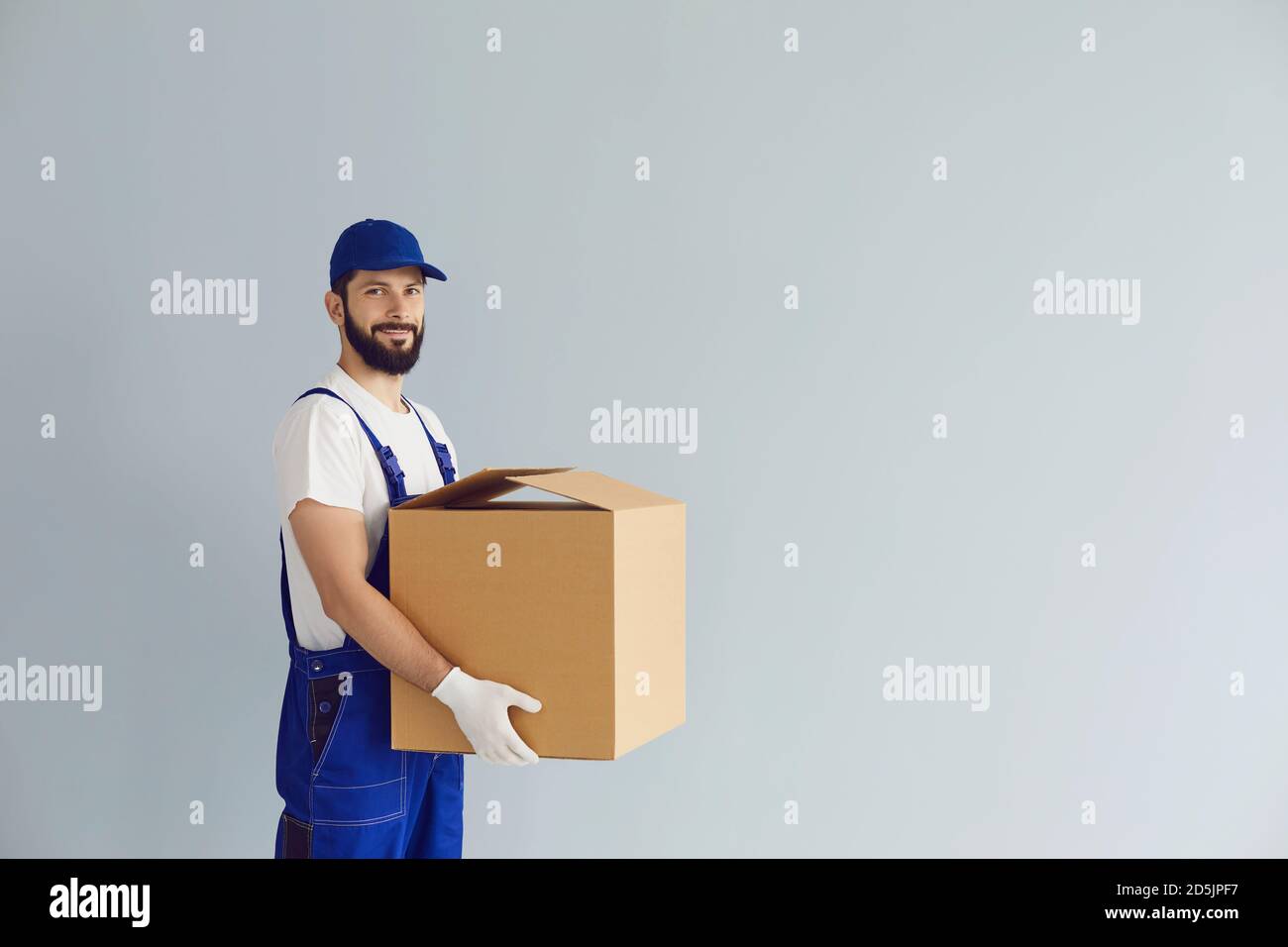 Lächelnder Lieferer hält Pappkarton in den Händen und schaut Bei der Kamera Stockfoto