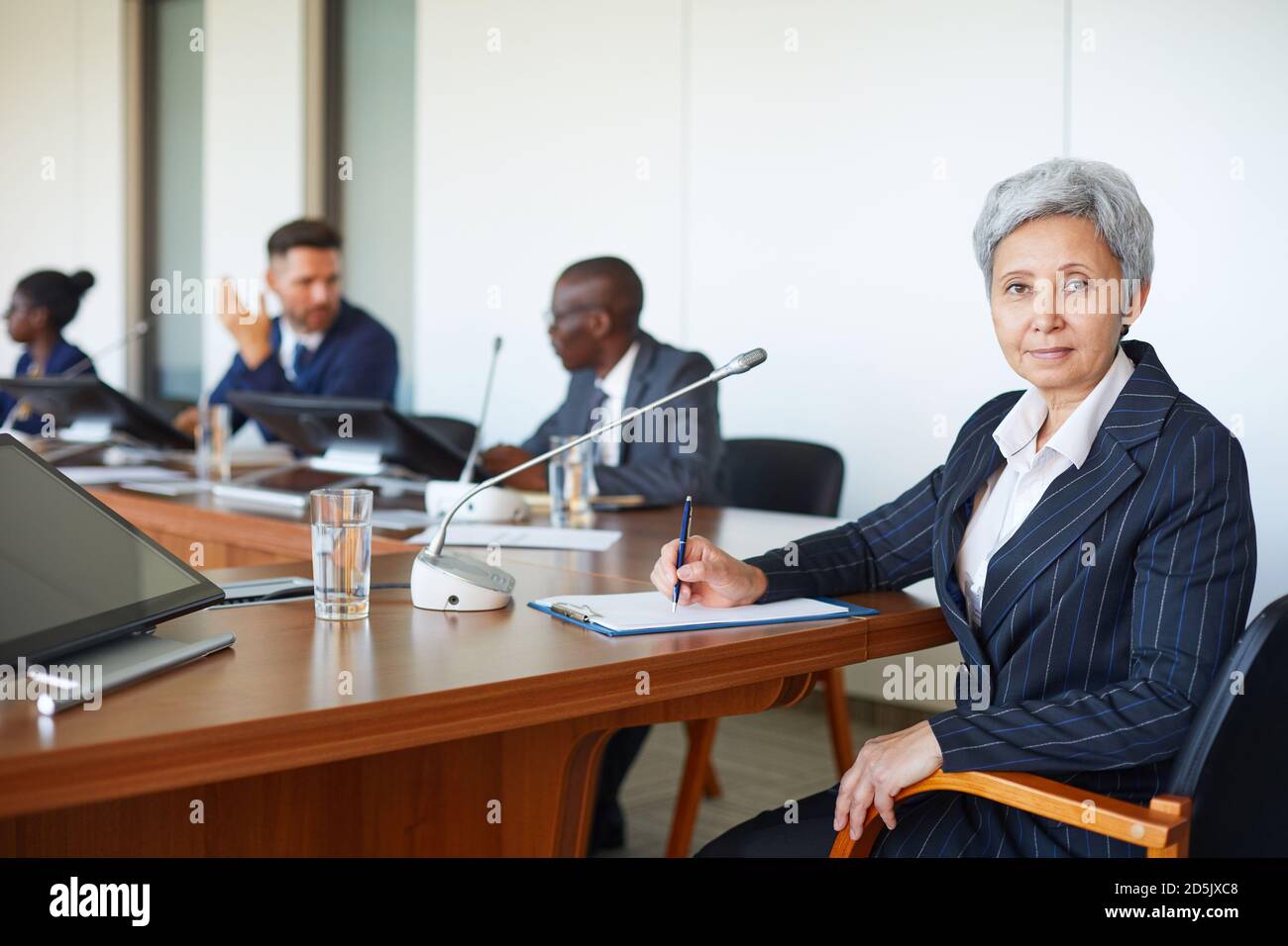 Porträt einer reifen Geschäftsfrau, die beim Sitzen auf die Kamera schaut Konferenz mit ihren Kollegen im Hintergrund Stockfoto