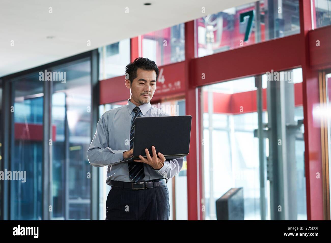 asiatischer Geschäftsmann, der in der Firmenlobby steht und E-Mails mit einem Laptop überprüft Computer Stockfoto