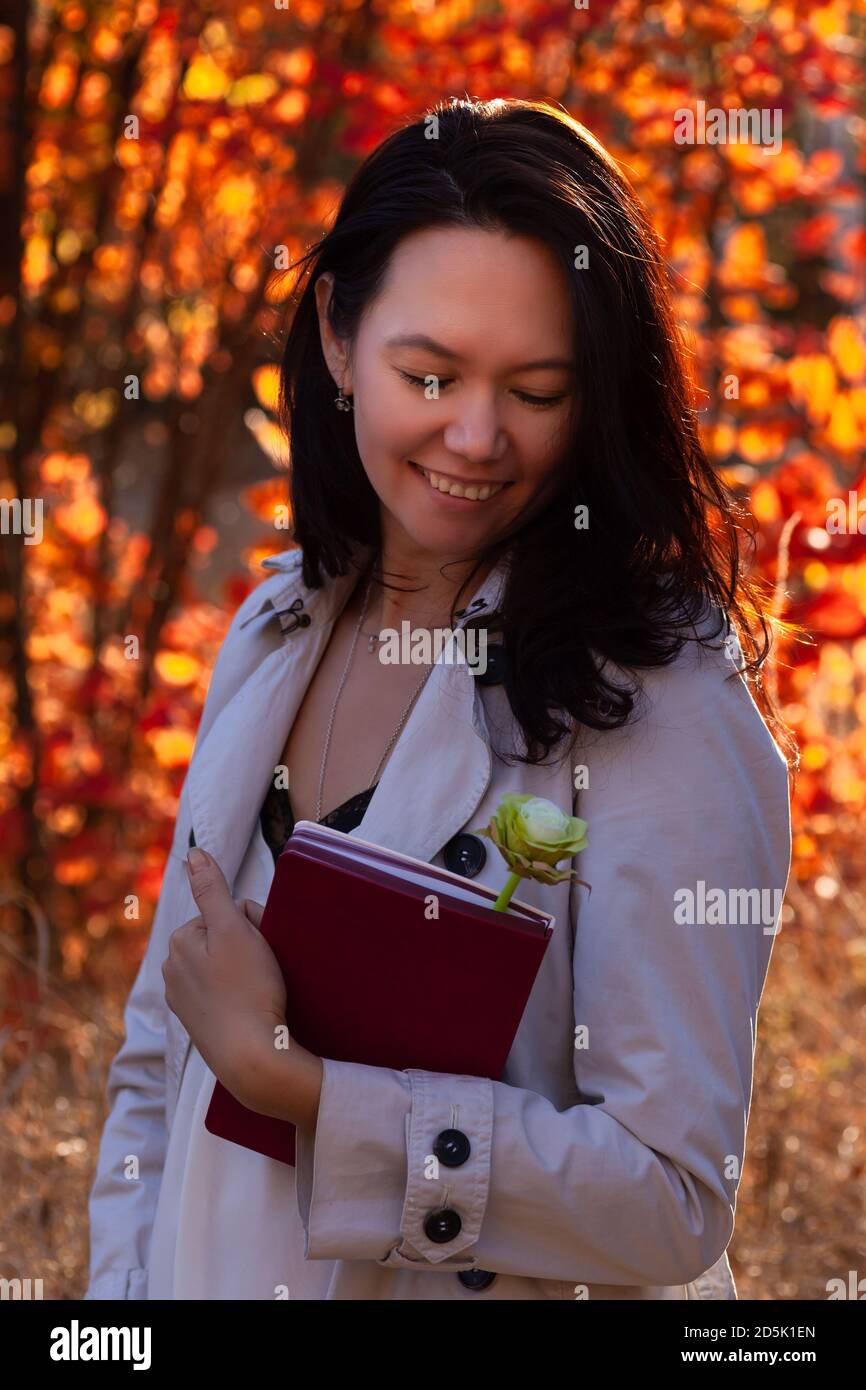 Portrait von schönen lächelnden asiatischen jungen Frau mit Buch und Blume im Herbstwald bei Sonnenuntergang Stockfoto