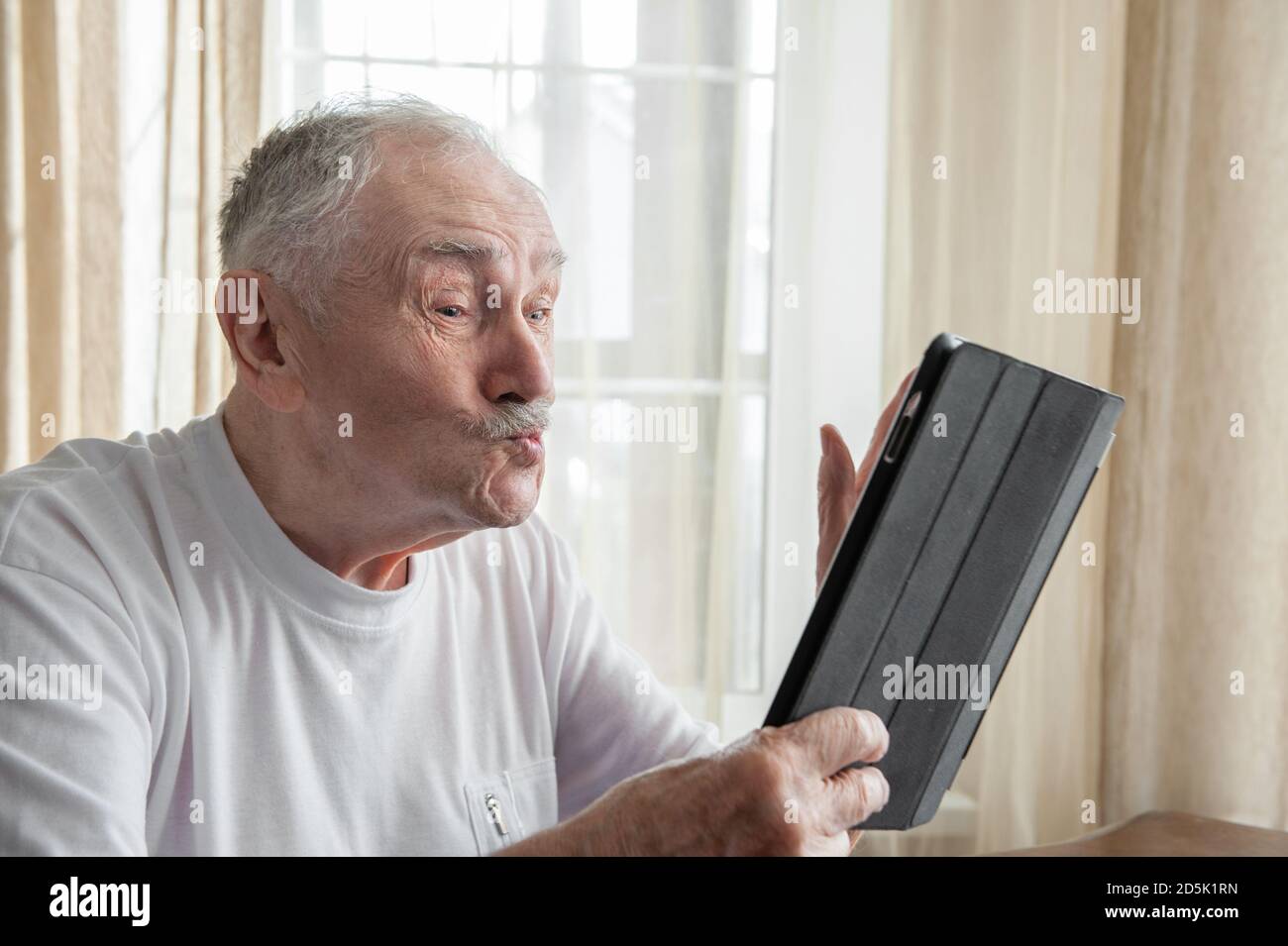 Zärtlich, grauhaarige, gutaussehende Mann in einem weißen T-Shirt bläst einen Kuss, während sie auf den Monitor des Gadgets.Konzepte der Kommunikation und Einsamkeit der Stockfoto