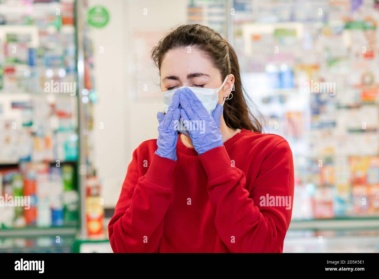 Eine junge Frau in einer medizinischen Maske und Handschuhen niest und bedeckt ihr Gesicht. Im Hintergrund der Shop Fenster von Apothekenprodukten. Krankheitsbild, V Stockfoto