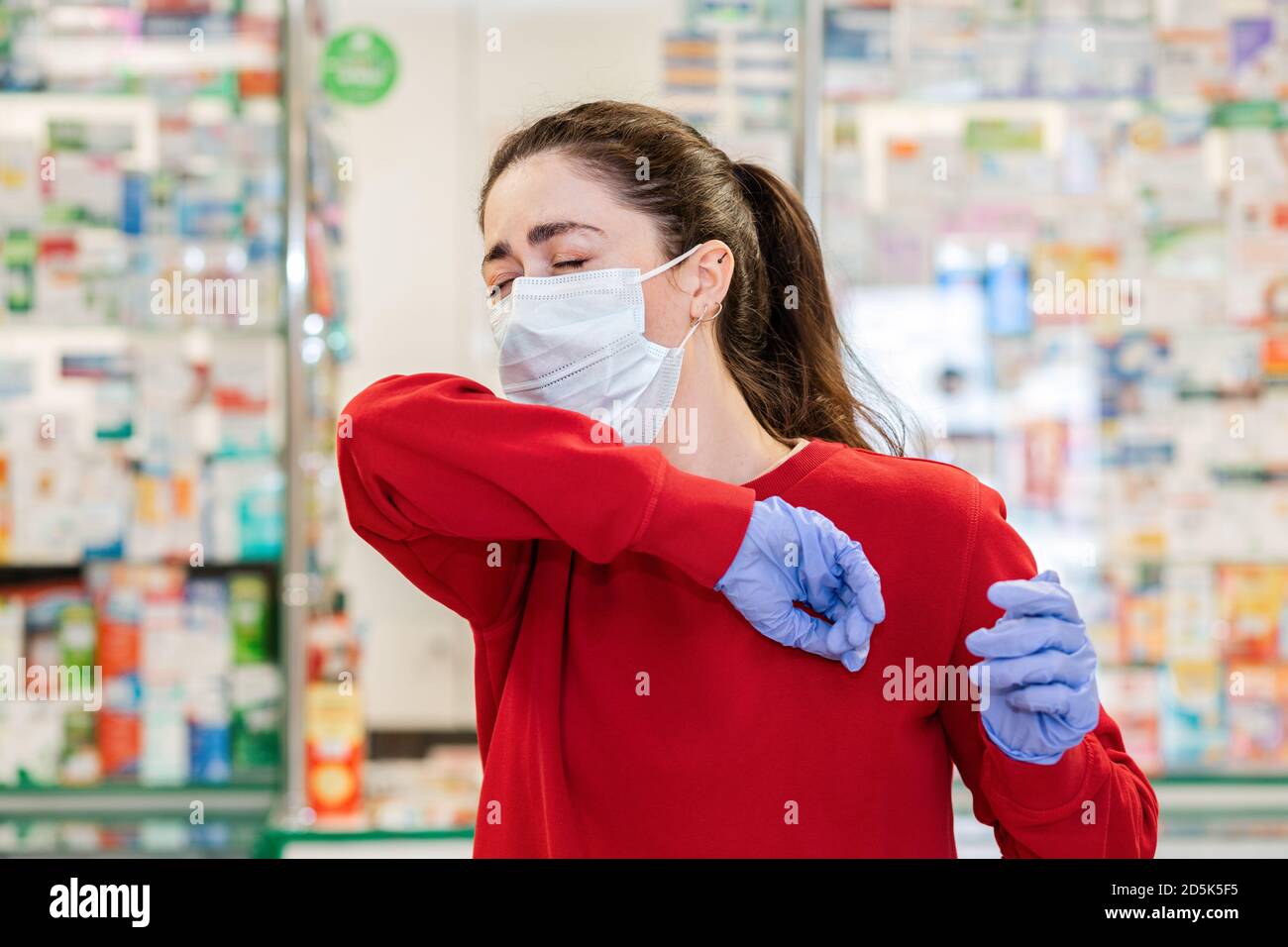 Eine junge Frau in einer medizinischen Maske und Handschuhen hustet und bedeckt ihr Gesicht mit der Ellenbogenbeuge. Im Hintergrund der Shop Fenster der Apotheke prod Stockfoto