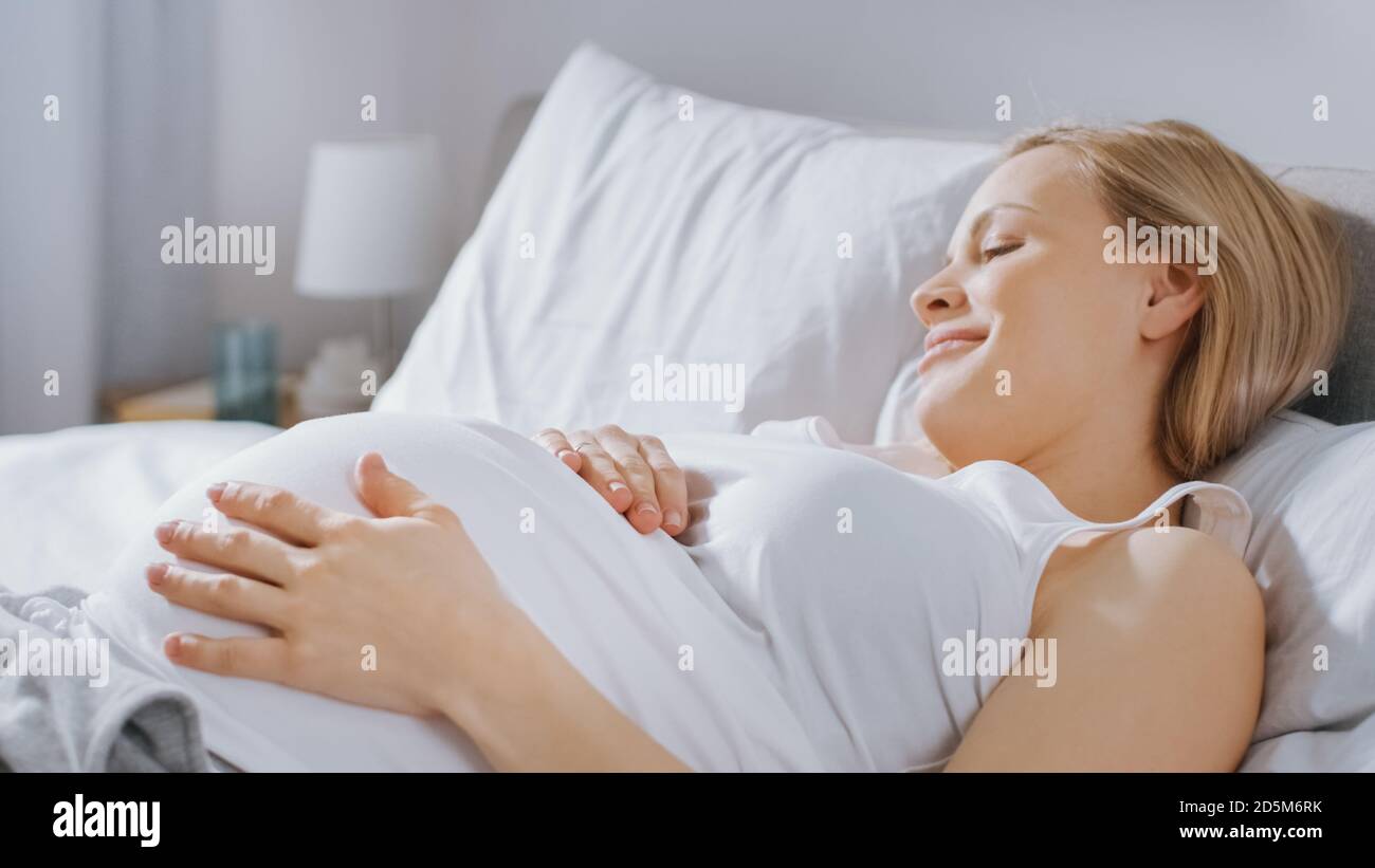 Glückliche junge schwangere Frau, die morgens im Bett liegt, berührt und streichelt sie ihren Bauch zärtlich, liebt ihr zukünftiges Baby und jeden Moment von ihr Stockfoto
