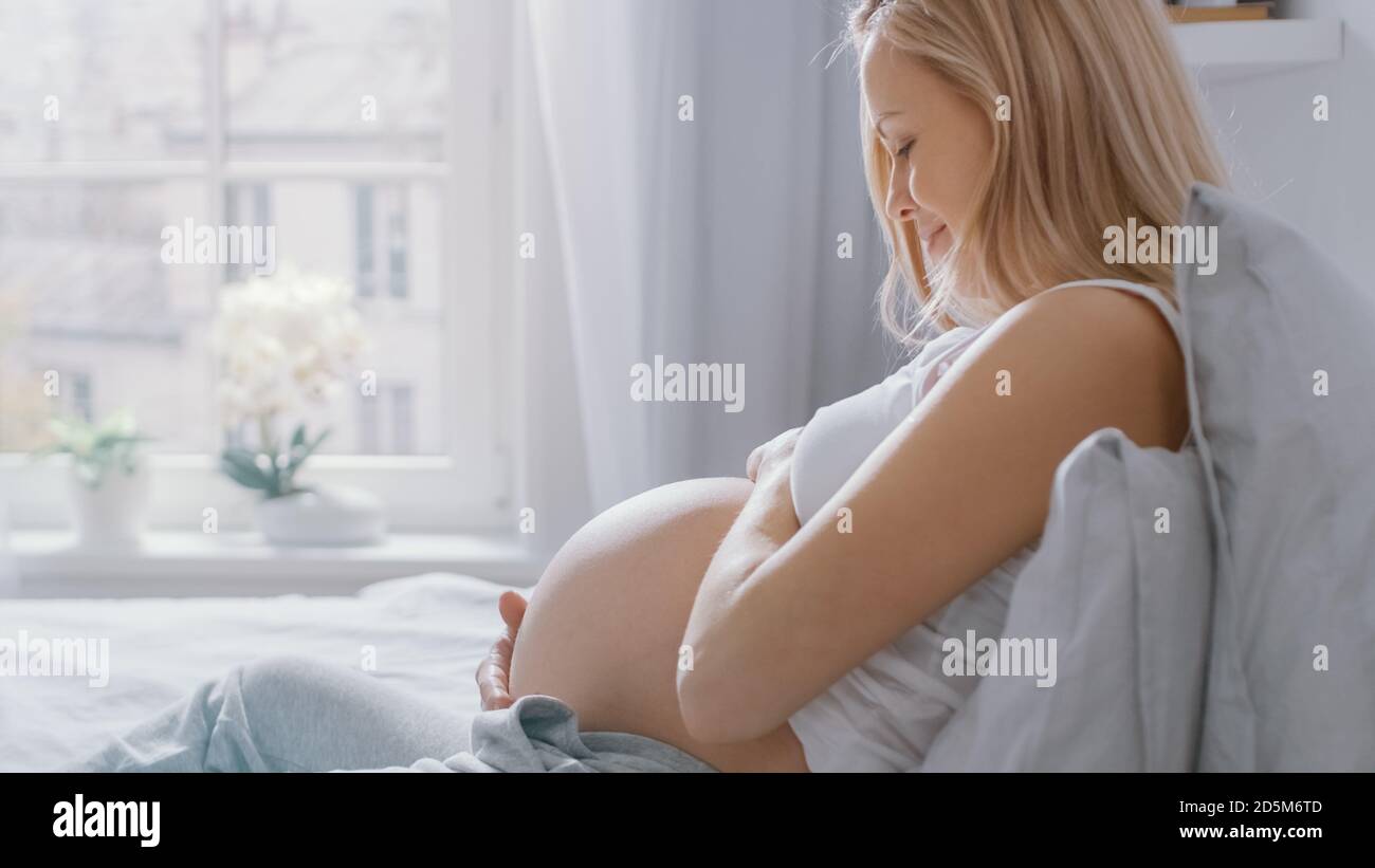 Glückliche junge schwangere Frau, die morgens im Bett liegt, berührt und streichelt sie ihren Bauch zärtlich, liebt ihr zukünftiges Baby und jeden Moment von ihr Stockfoto