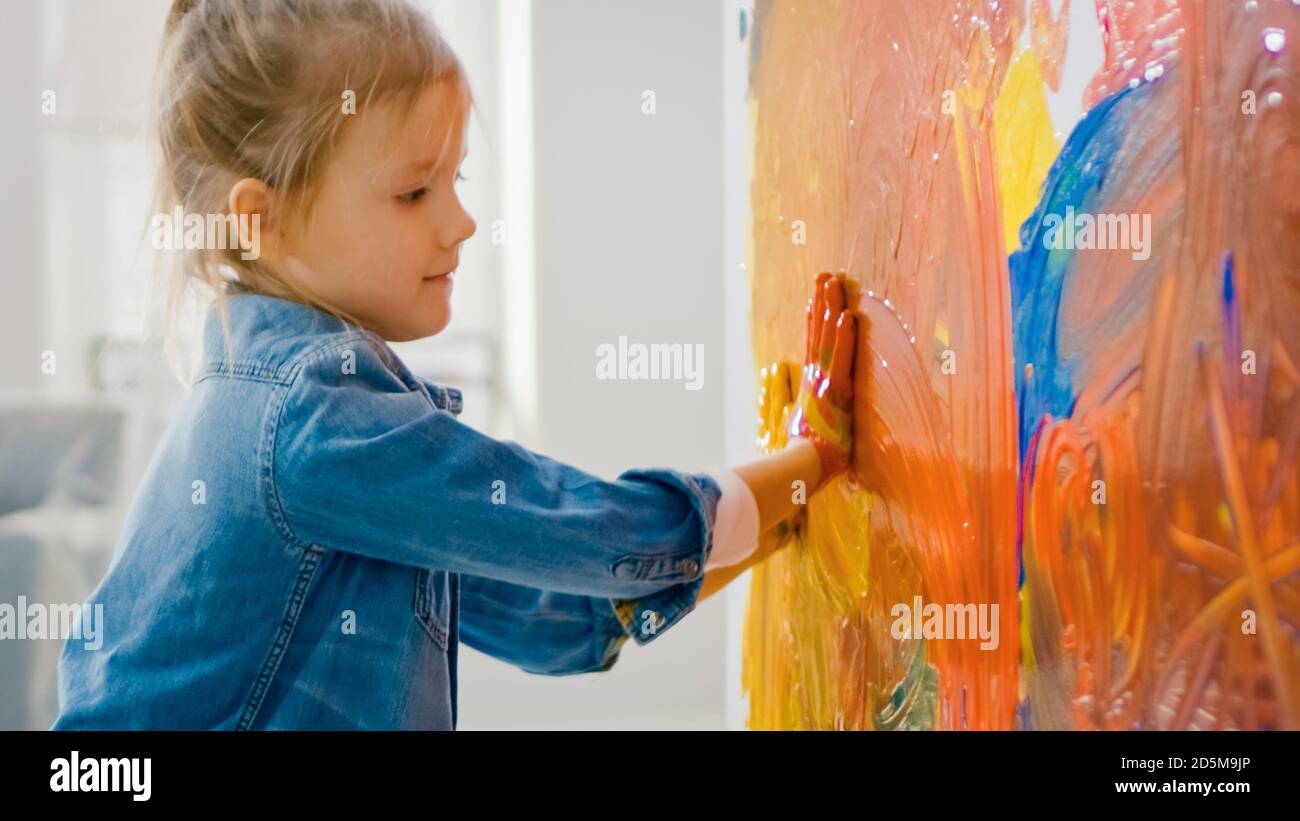 Happy Little Girl mit Händen getaucht in Vivid Paint zeichnet bunte Abstraktionen an der Wand. Sie hat Spaß und lacht. Das Haus wird renoviert. Stockfoto
