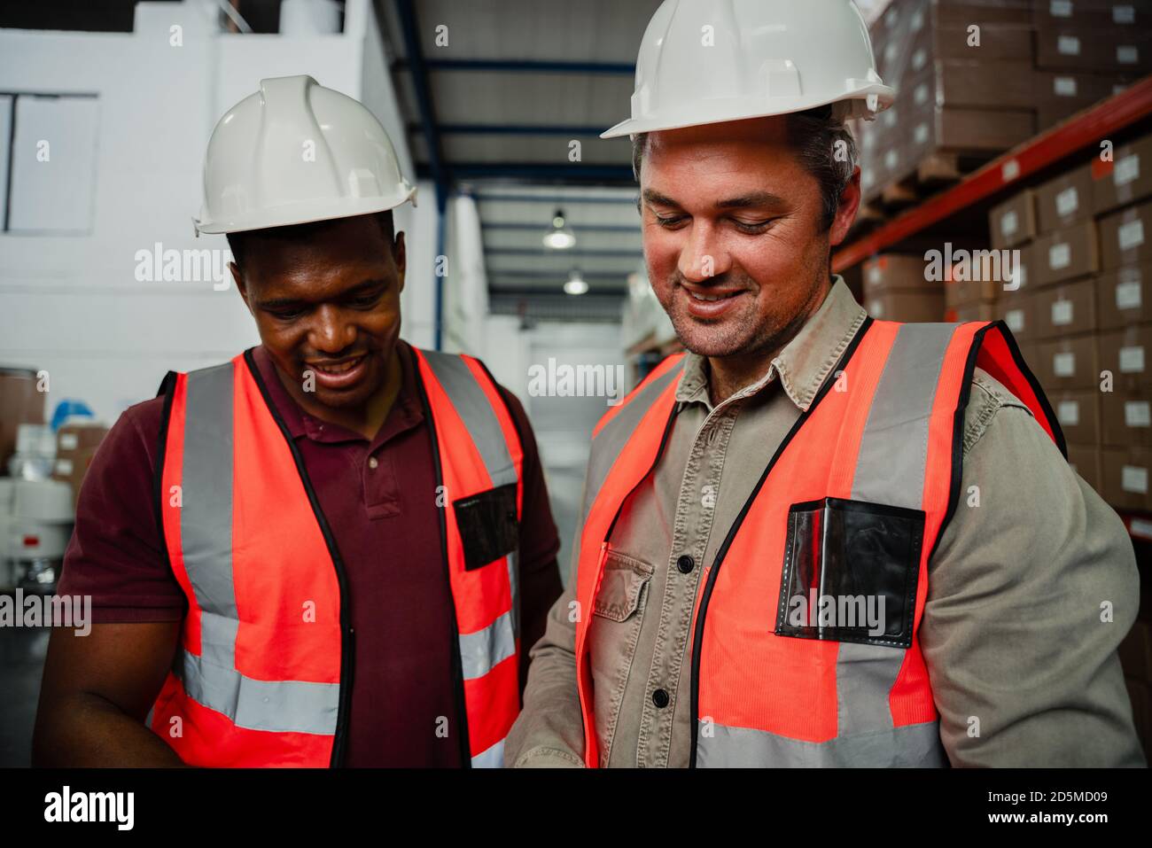 Lächelnde Kollegen tragen Overalls und Hüte, die in der Fabrik lachen Stockfoto