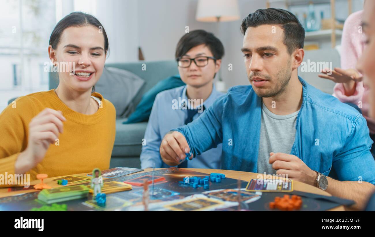 Diverse Gruppe von Jungs und Mädchen spielen in einem strategischen Brettspiel mit Karten und Würfel. Die Spieler Des Teams Machen Die Fünf Höchsten Punkte. Gemütliches Wohnzimmer in einem Tag Stockfoto