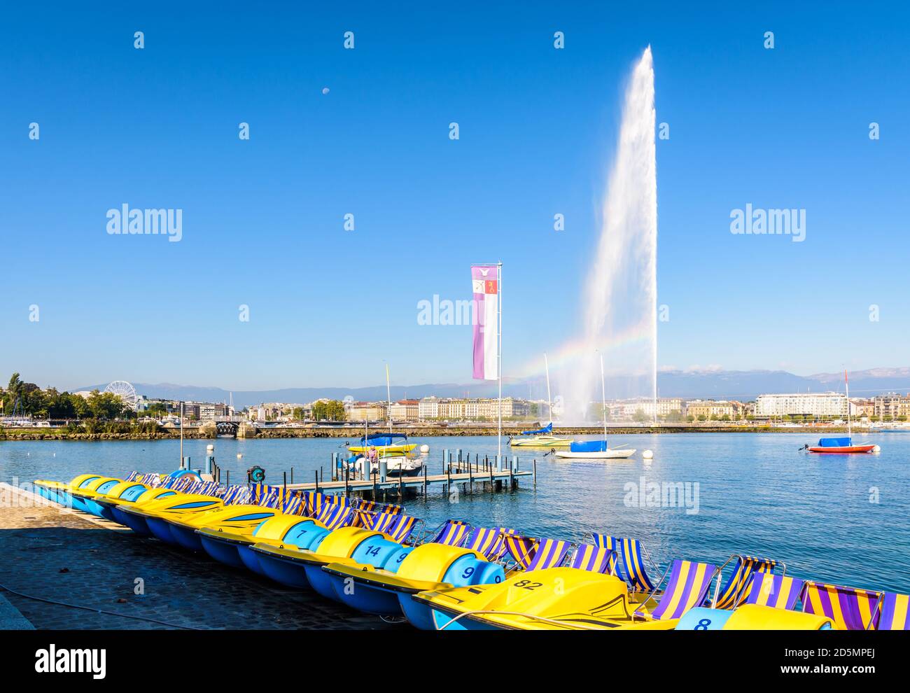 Tretboote Reihen sich an einem sonnigen Sommermorgen am Ufer des Genfer Sees mit dem Jet d'Eau Wasserstrahlbrunnen in der Ferne an. Stockfoto
