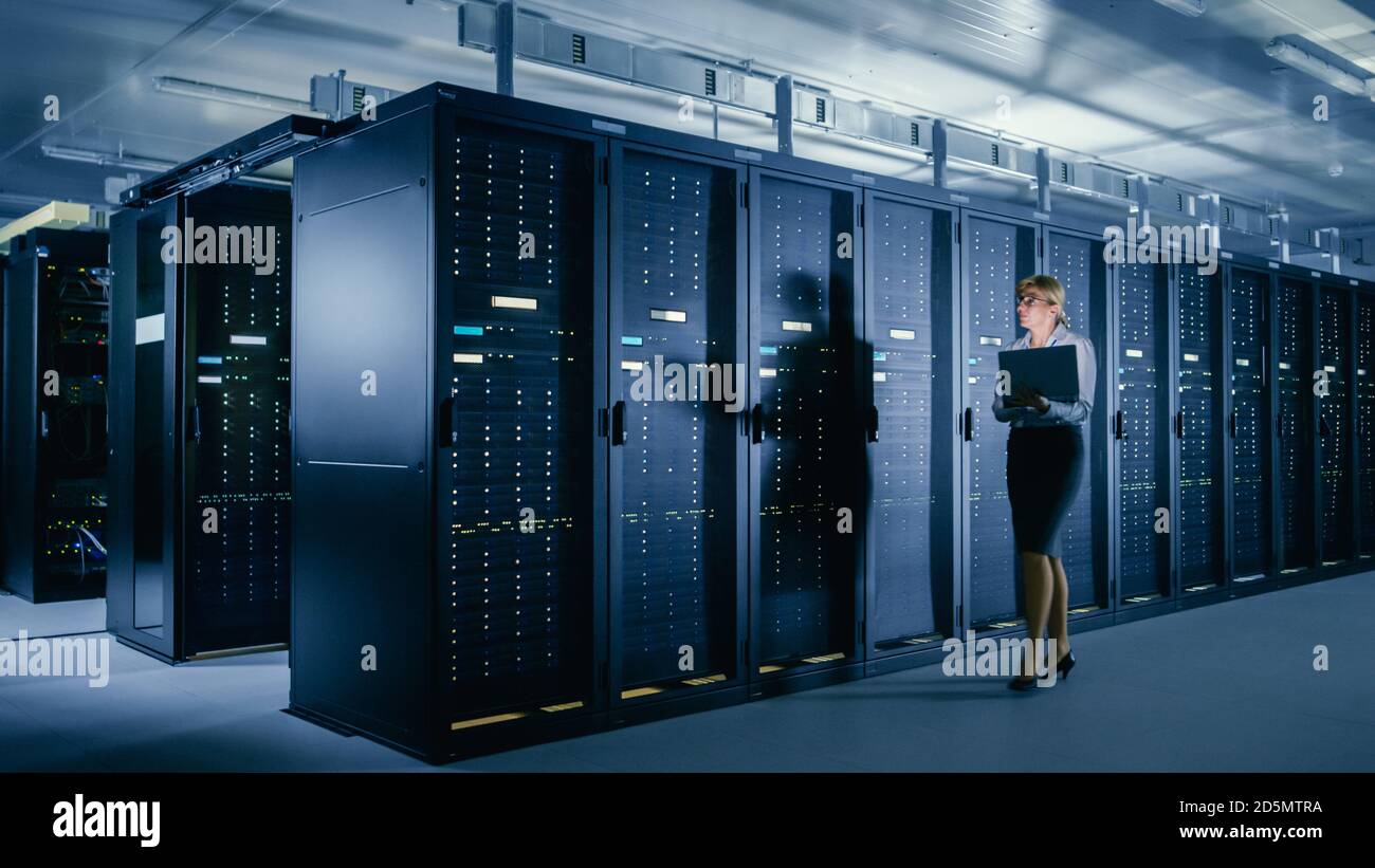 Im Rechenzentrum: Weibliche IT-Technikerin läuft neben Reihe von Server-Racks, verwendet Laptop-Computer, um Wartungsdiagnose auszuführen, damit Mainframe funktioniert Stockfoto