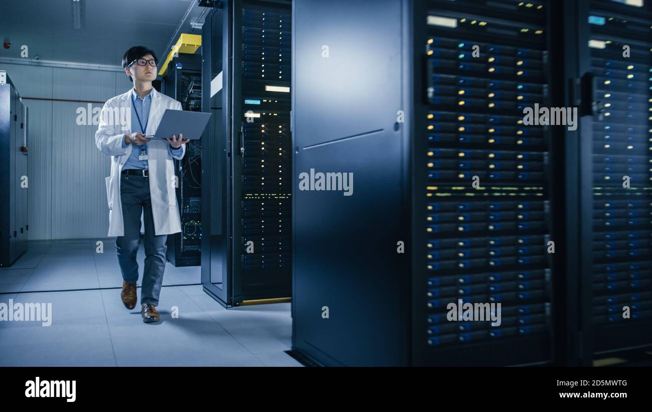 Im Rechenzentrum: Männlicher IT-Techniker, der White Coat trägt und neben Server-Racks läuft, verwendet Laptop-Computer, um Wartungs-Diagnose-Tools auszuführen. Er ist es Stockfoto