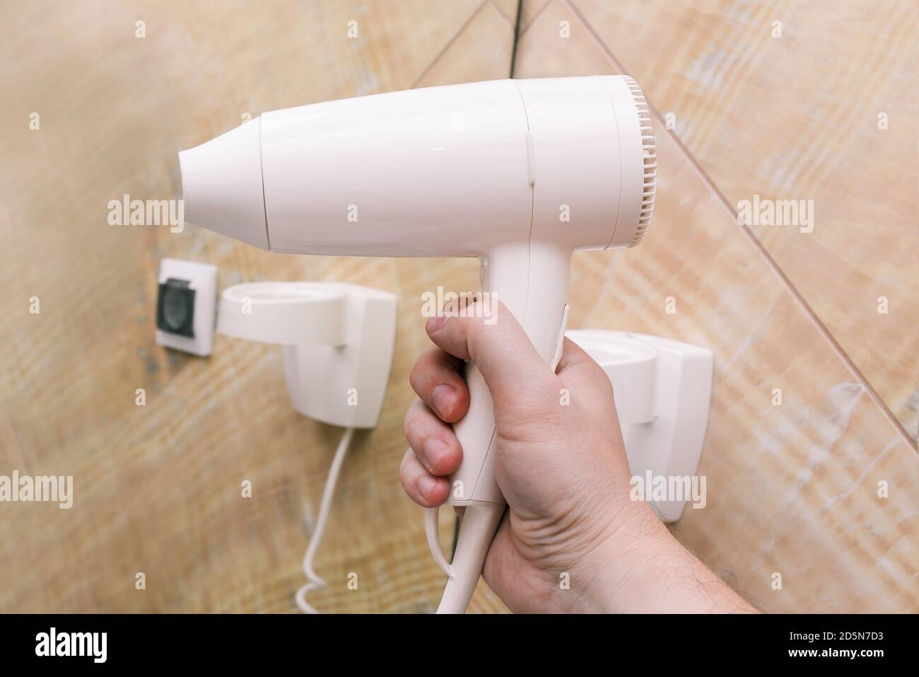 Weißer Haartrockner in Nahaufnahme. Ein Mann hält einen Plastiktrockner in der Hand vor einem Hintergrund aus Spiegeln und Marmorfliesen. Interieur eines Badezimmers, Schönheit Stockfoto