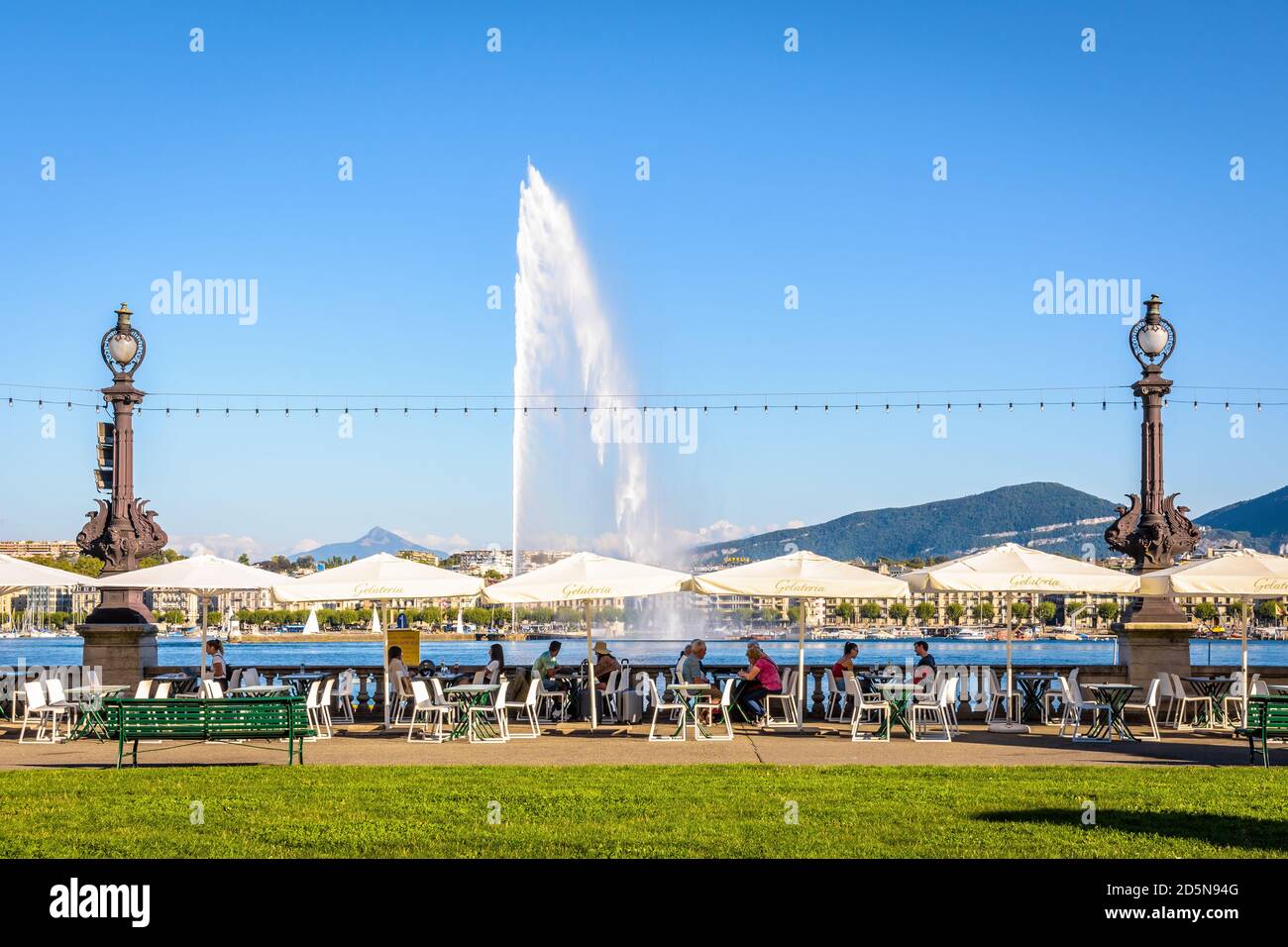 Touristen genießen die sonnige Terrasse einer Eisdiele an der Bucht von Genf, mit Panoramablick über den Wasserstrahlbrunnen und den Mont Blanc. Stockfoto