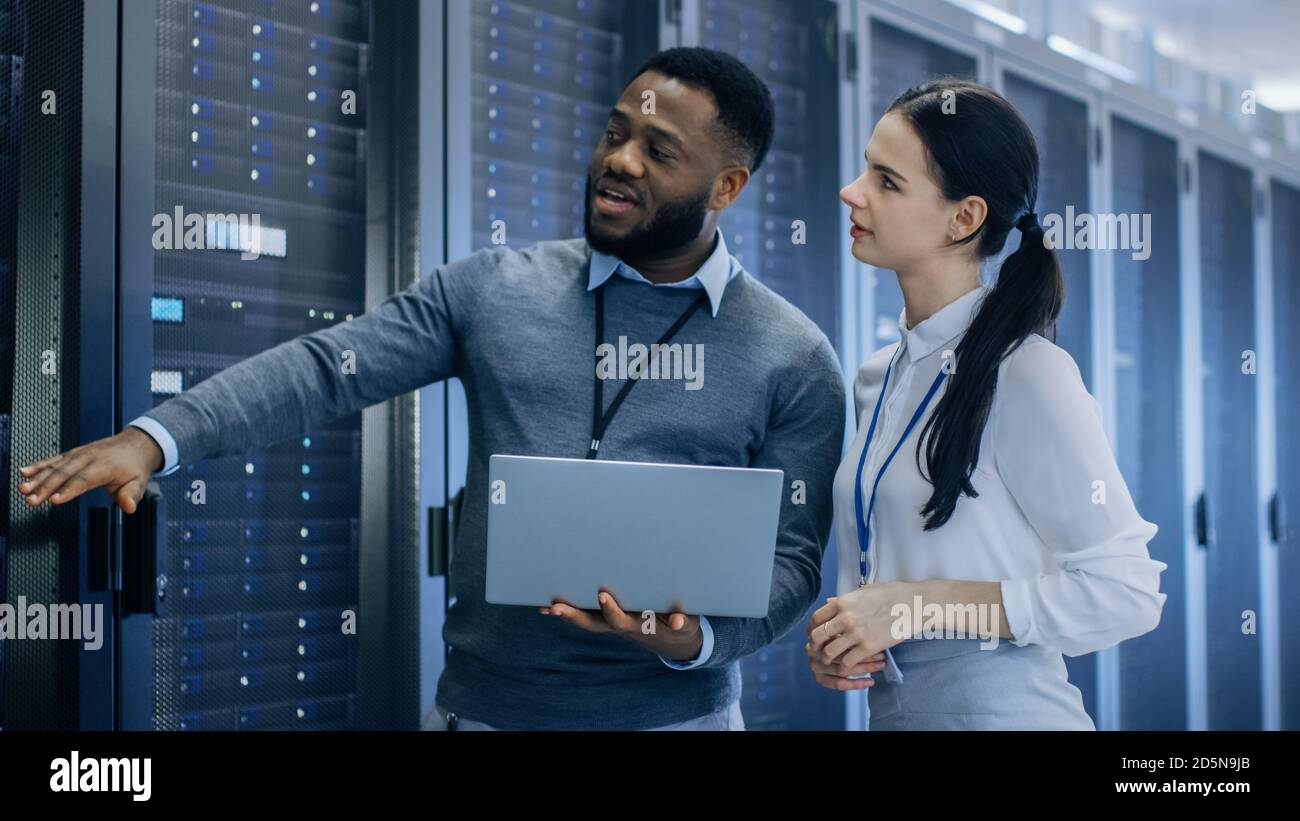 Schwarzer IT-Techniker mit Laptop gibt einem jungen Praktikant eine Tour. Sie sprechen im Rechenzentrum, während sie neben Server-Racks gehen. Läuft Stockfoto