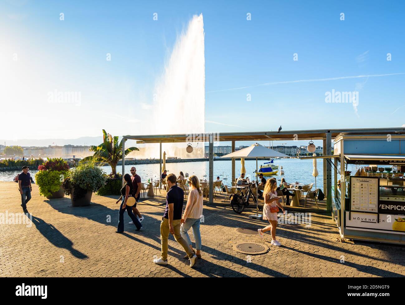 Menschen, die am Ende eines sonnigen Tages am Genfer See spazieren oder die Terrasse einer Eisdiele mit Blick auf den Wasserstrahlbrunnen genießen. Stockfoto