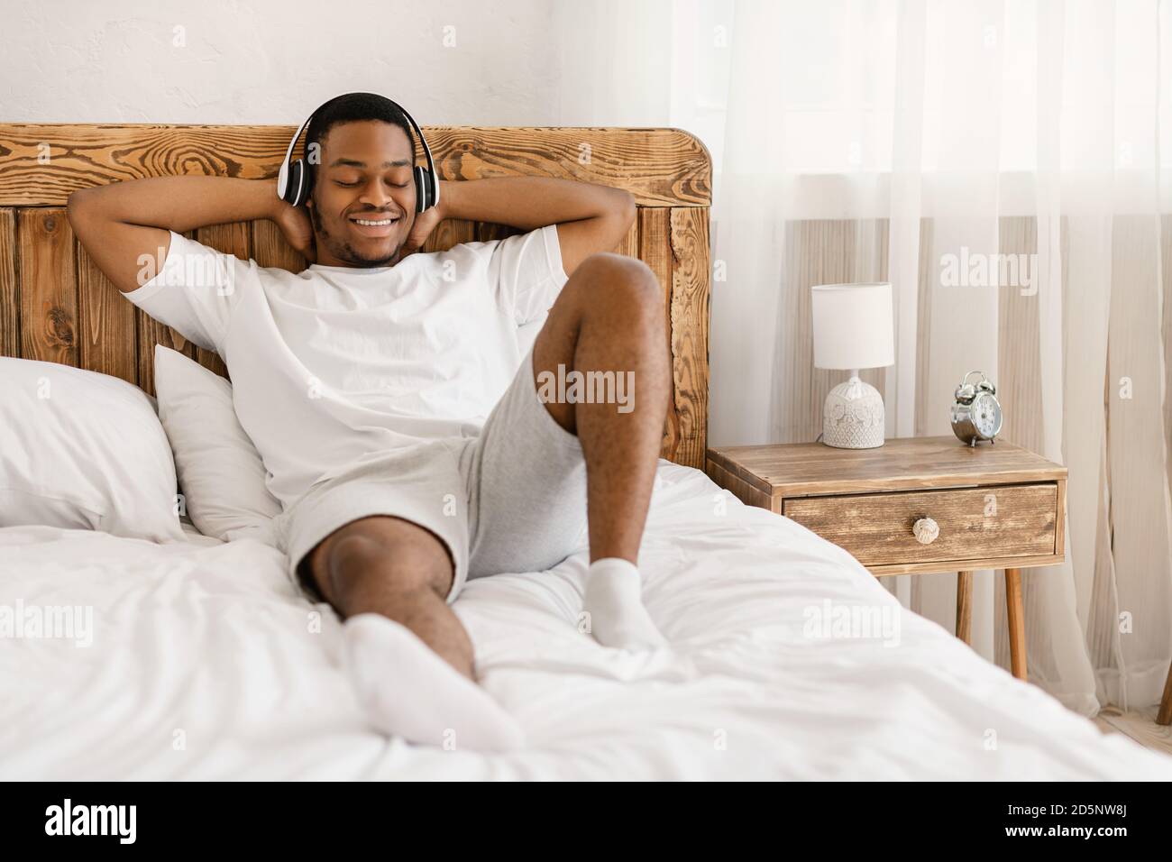 Entspannter Afrikanischer Mann Musik In Den Kopfhörern Im Bett Drinnen Hören Stockfoto