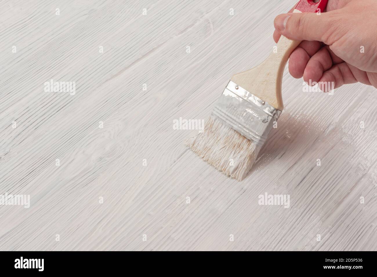 Gebürstete Holzoberfläche mit weißer Farbe mit einem Pinsel bemalt In einer männlichen Hand Stockfoto